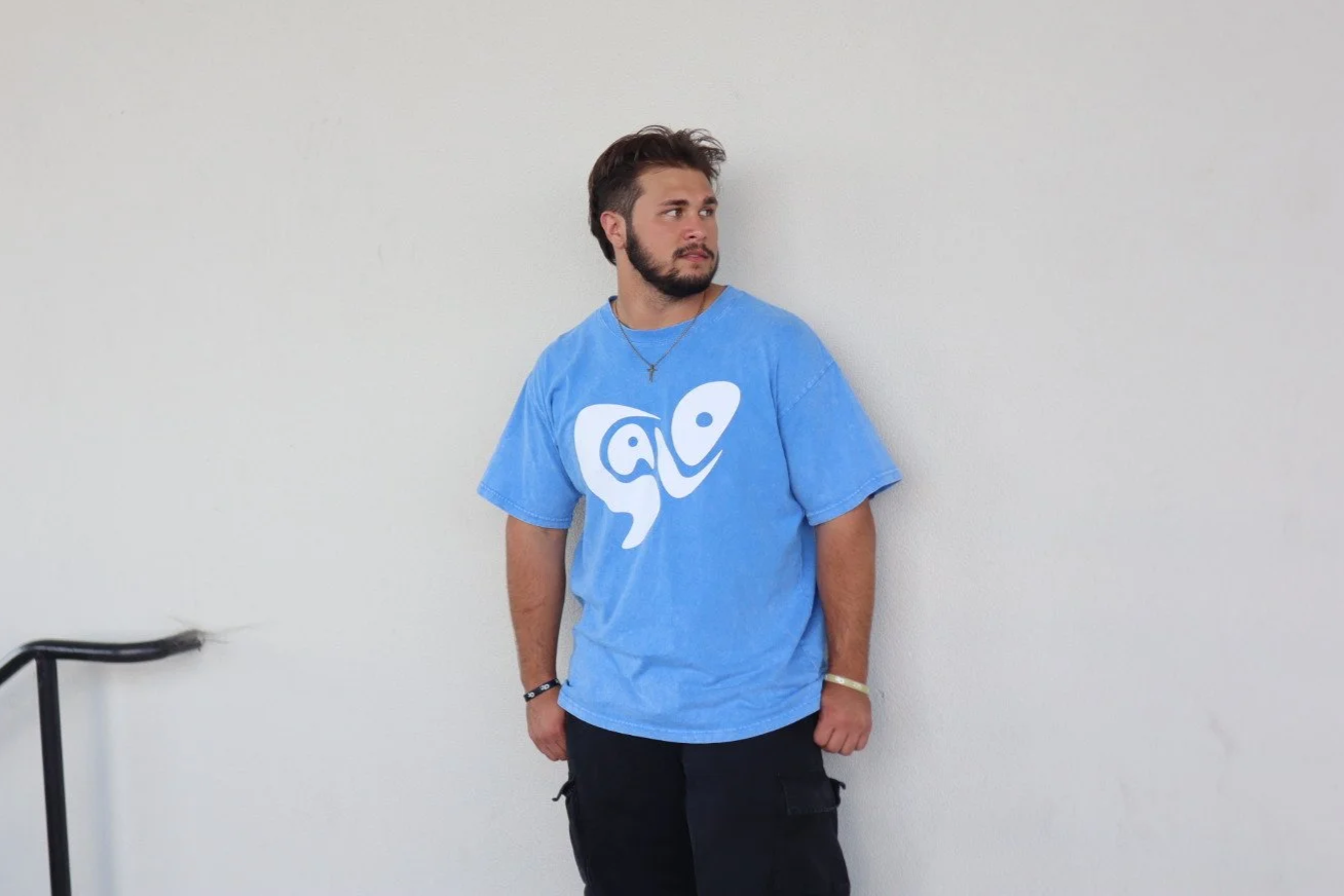 A young man with a beard and brown hair, wearing a blue graphic t-shirt, standing against a plain white wall.