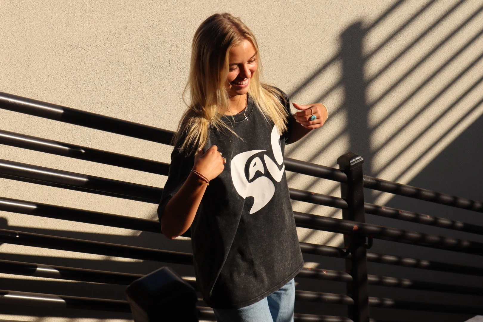 A young woman with blonde hair smiling outdoors at sunset, wearing a black T-shirt with a white logo, light blue jeans, and jewelry, standing next to a metal railing with shadows cast on a beige wall.