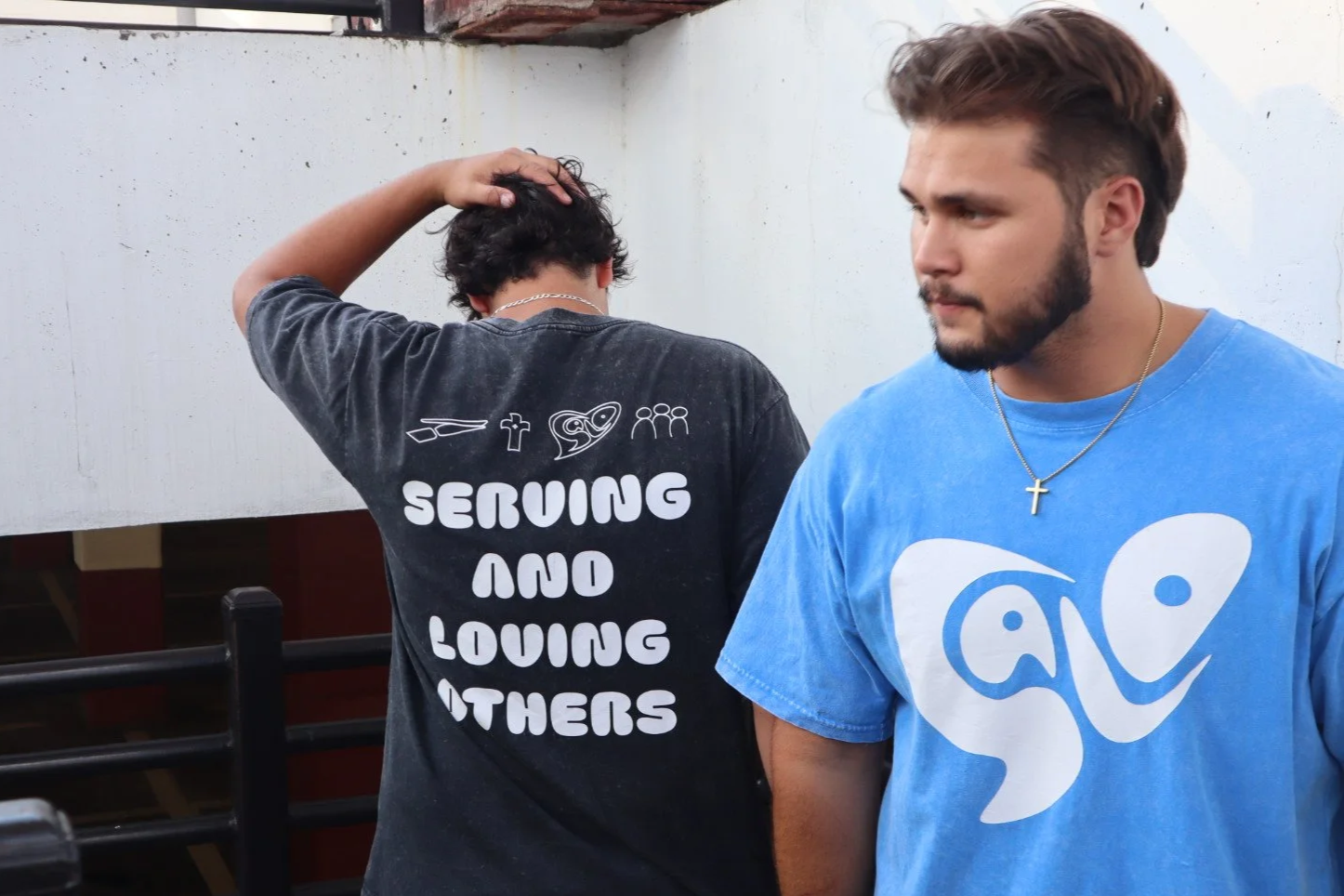 Two young men standing outdoors against a white wall; one has his hand on his head, wearing a black T-shirt with religious and love symbols and the words "Serving and Loving Others," and the other is looking to the side, wearing a blue T-shirt with a white abstract design and a gold cross necklace.