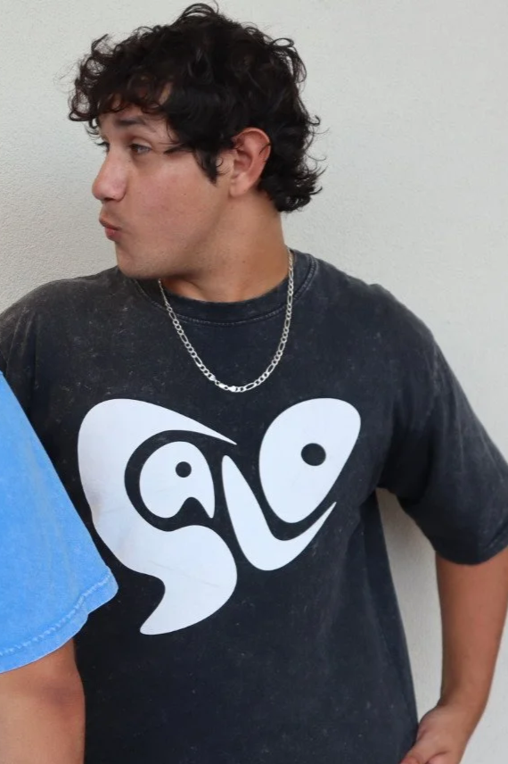 A young man with curly dark hair, wearing a black T-shirt with a large white abstract face design, and a silver chain necklace, standing against a plain light-colored wall.
