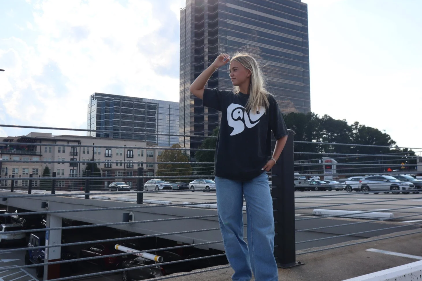 A young woman with blonde hair standing outdoors on a parking lot with tall buildings in the background. She is wearing a black t-shirt with a white graphic and light blue jeans, and is looking to the side while holding her hand to her forehead.