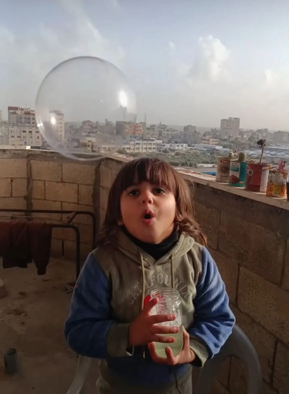 A young child with brown hair, wearing a jacket, blowing a bubble using soap and water, holding a jar of soapy solution. The background shows Gaza city skyline with buildings and a cloudy sky.