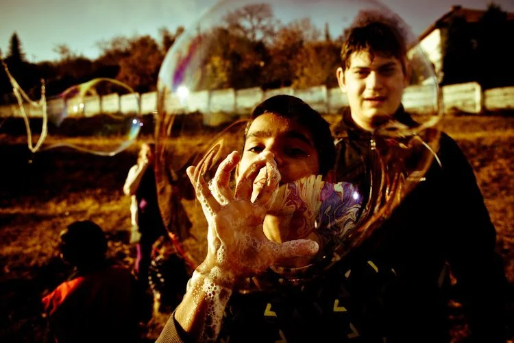 Bubbles Not Bombs and a child making soap bubbles using his hands.  He has soap suds on his arms, and looks happy and smiling in the sunshine of a refugee camp