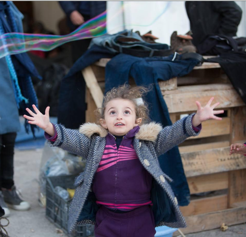 A young girl with curly hair and a coat reaching up with her arms open wide to play with bubbles.