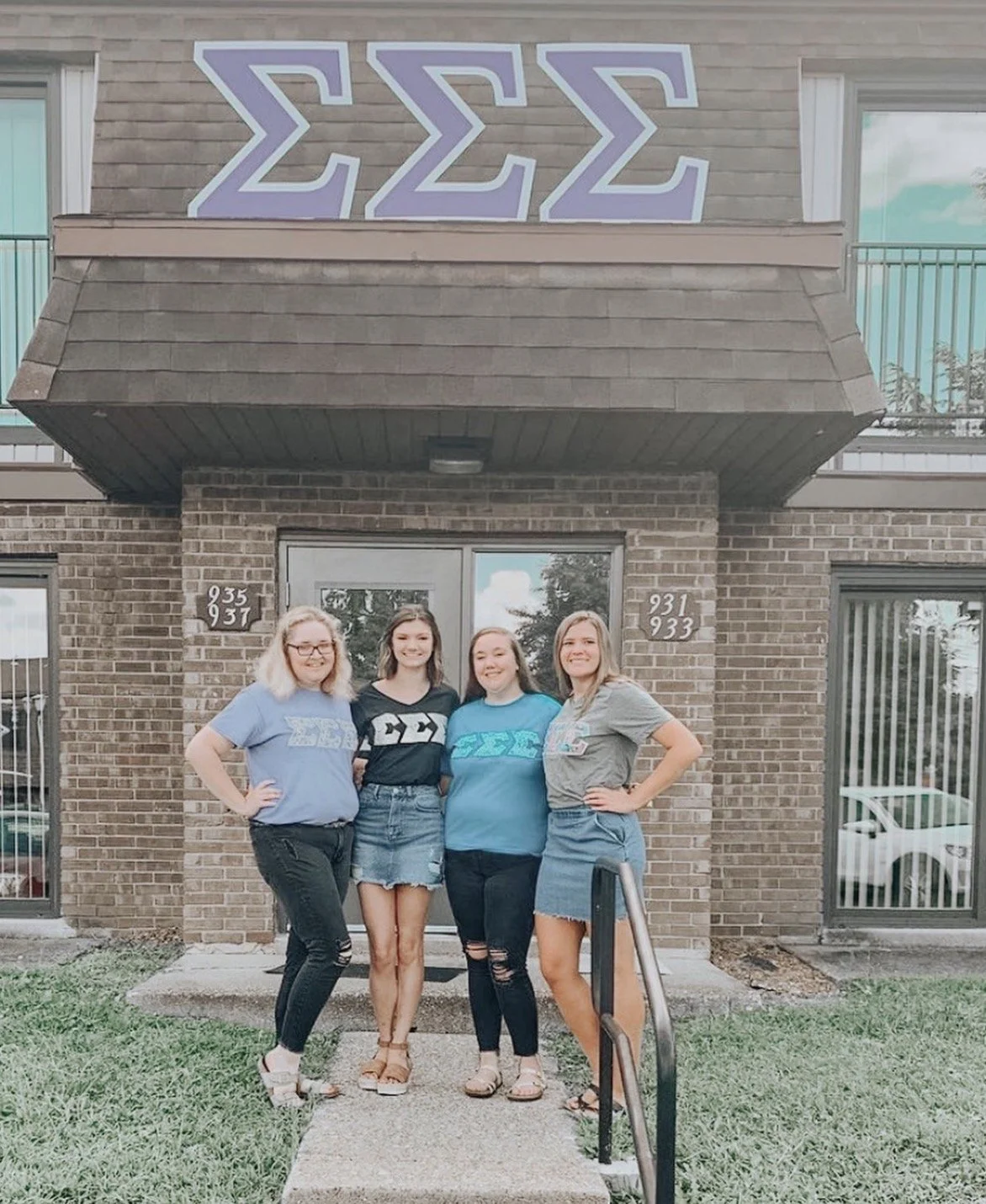 Four young women standing in front of a brick apartment building with the Greek letters Sigma Sigma Sigma on the wall above the entrance, all wearing sorority shirts.