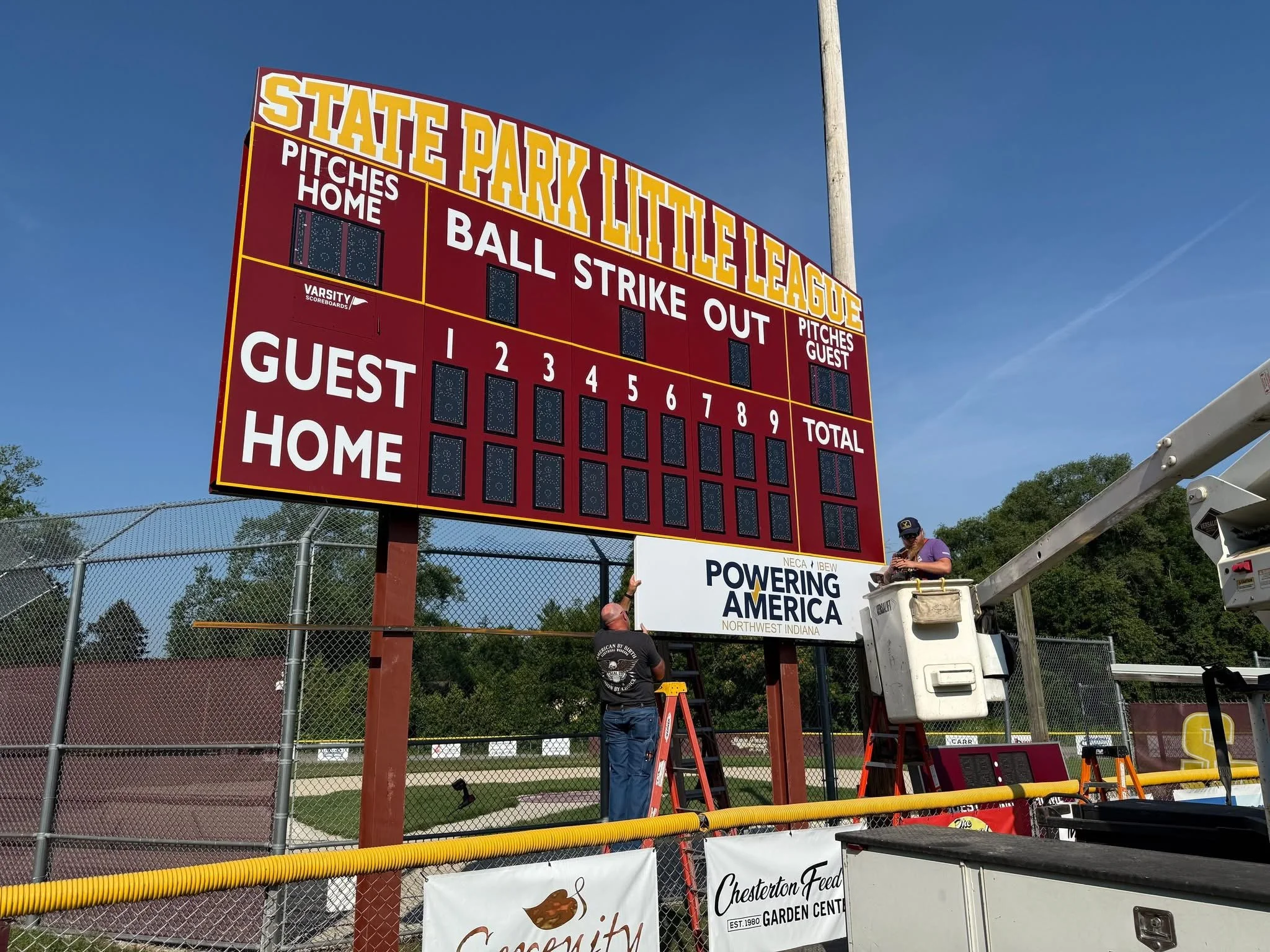 A large scoreboard at a baseball field with the title 'State Park Little League'. It has sections for 'Pitches Home', 'Ball Strike Out', 'Pitches Guest', and 'Guest Home', as well as spaces for tracking the count of balls and strikes. Two people are 