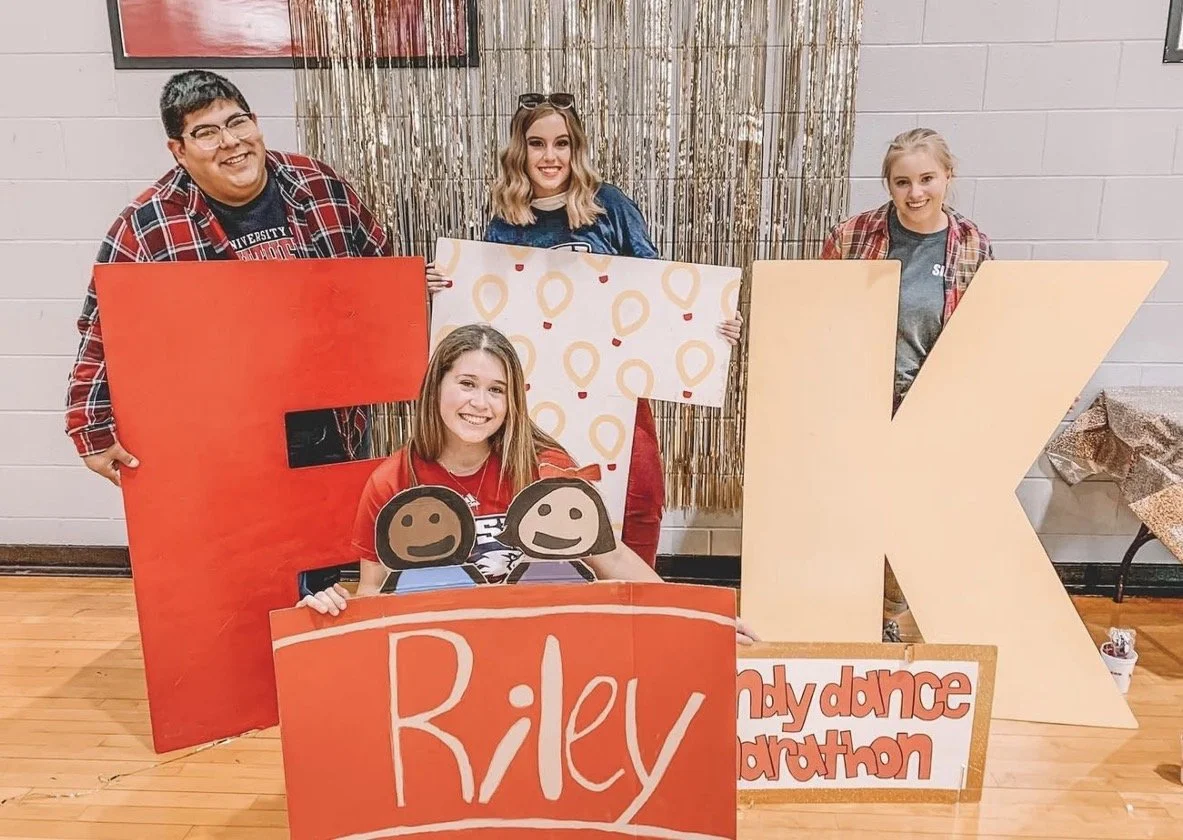 Five young women posing behind large cardboard letters spelling out "PEEK" at a relay for life event. They are smiling and holding colorful signs and cutouts, with a shiny gold curtain in the background.