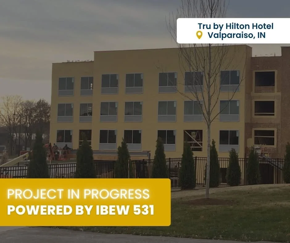 Photo of a multi-story building under construction at the Tru by Hilton Hotel in Valparaiso, Indiana, with a yellow banner stating "Project in Progress Powered by IBEW 531".
