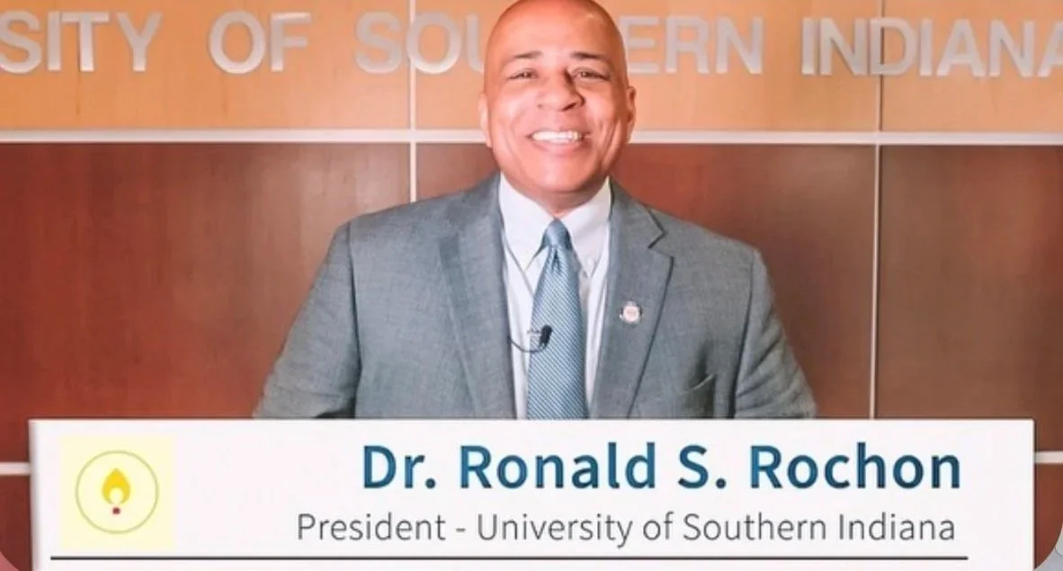 A smiling man in a gray suit and blue tie standing in front of a wooden wall with text "University of Southern Indiana" partly visible, with a presentation banner displaying "Dr. Ronald S. Rochon, President - University of Southern Indiana."