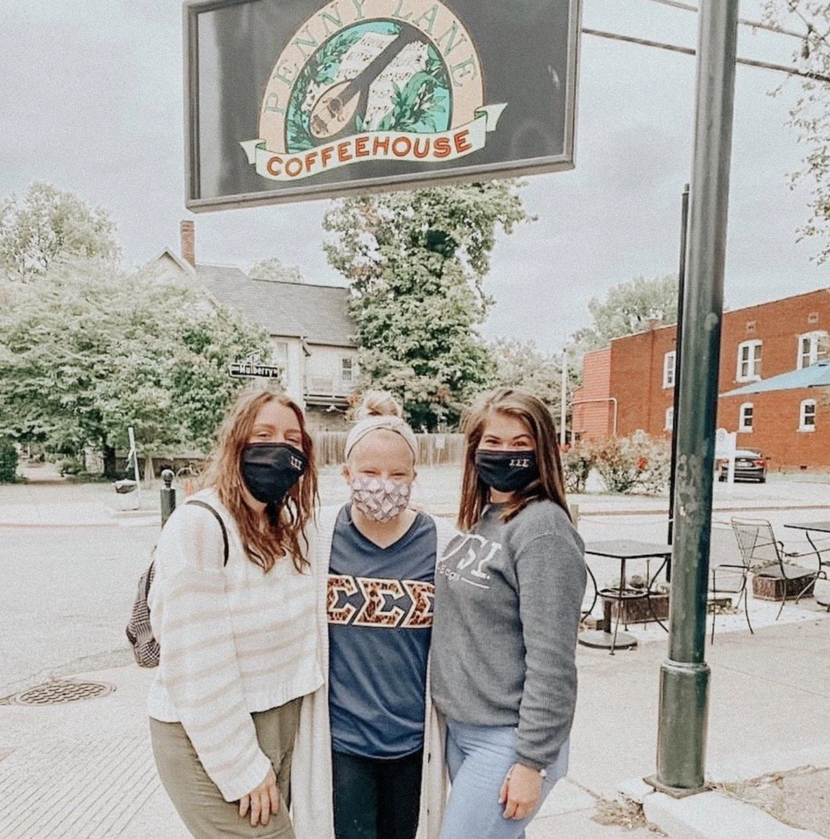 Three women standing outdoors in front of a coffeehouse sign, all wearing face masks, smiling for the camera.