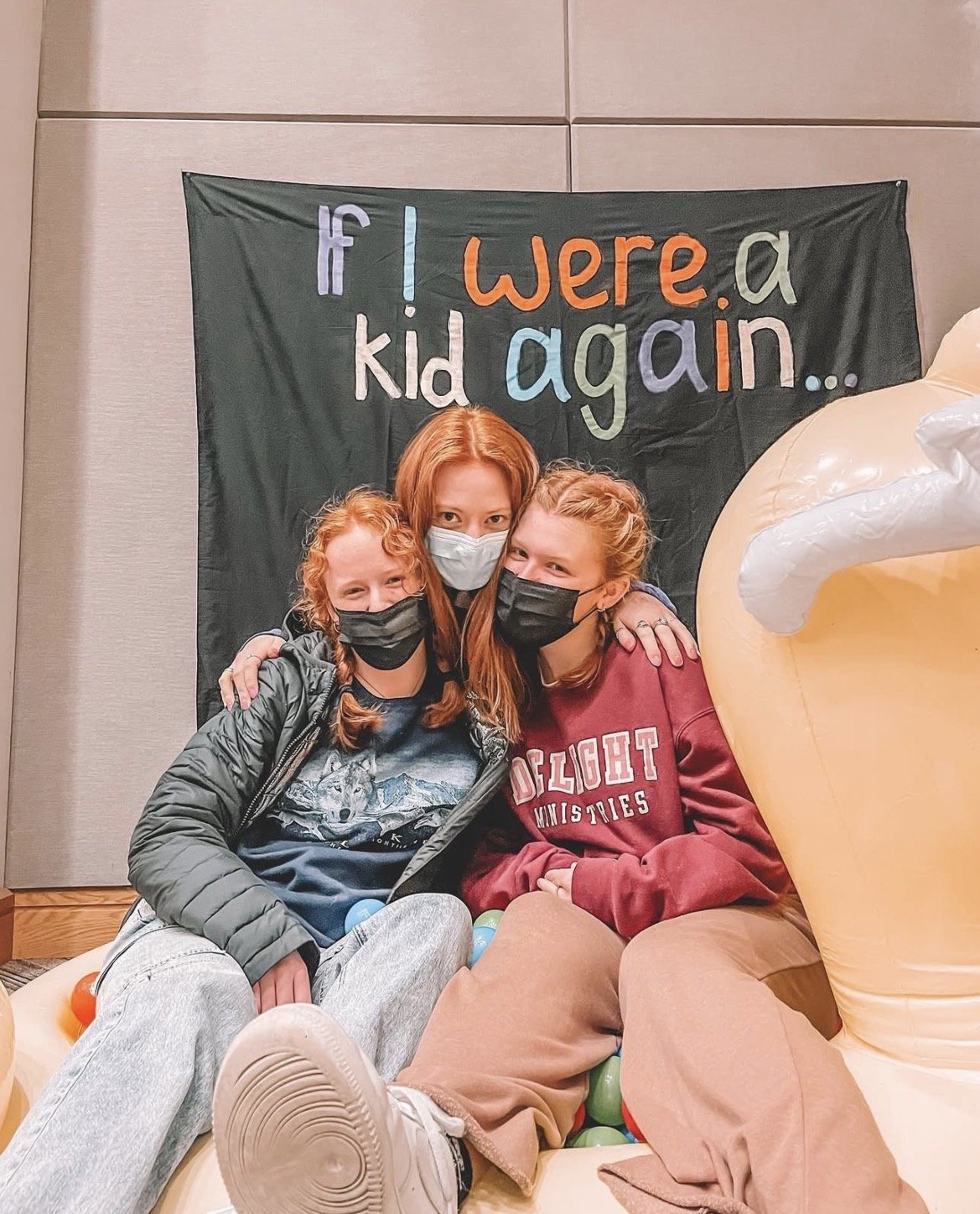 Three people sitting on a couch with a large inflatable figure partially visible on the right. All are wearing masks and smiling. Behind them is a green banner with colorful text that reads, 'if I were a kid again...'.
