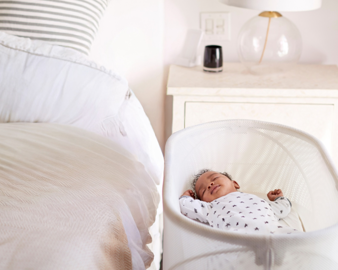 A sleeping baby in a white bassinet near a bed with white sheets and striped pillow