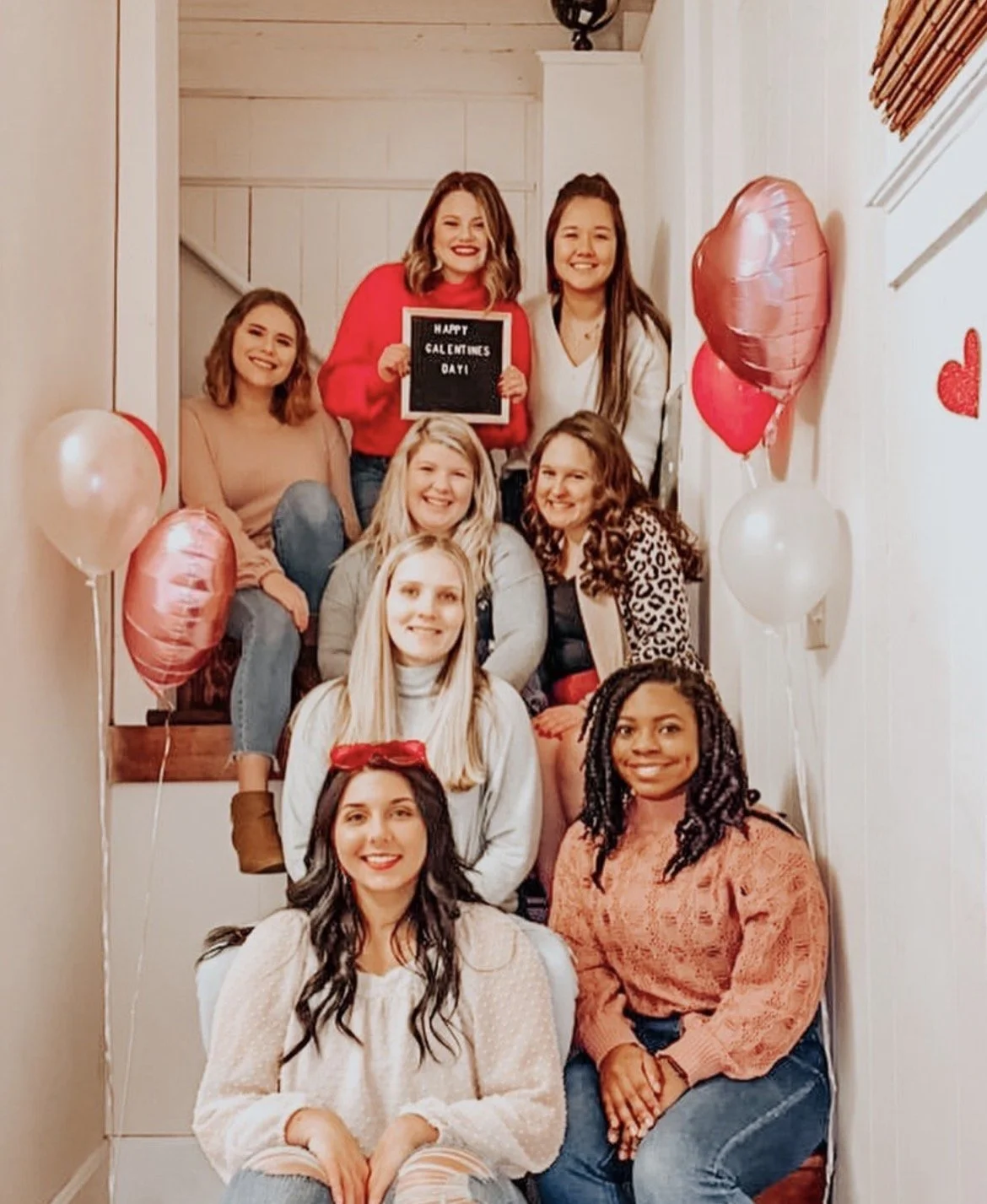 Group of seven smiling women sitting on staircase decorated with pink, red, and white balloons for Valentine's Day, one woman holding a sign that says 'Happy Valentine's Day'.