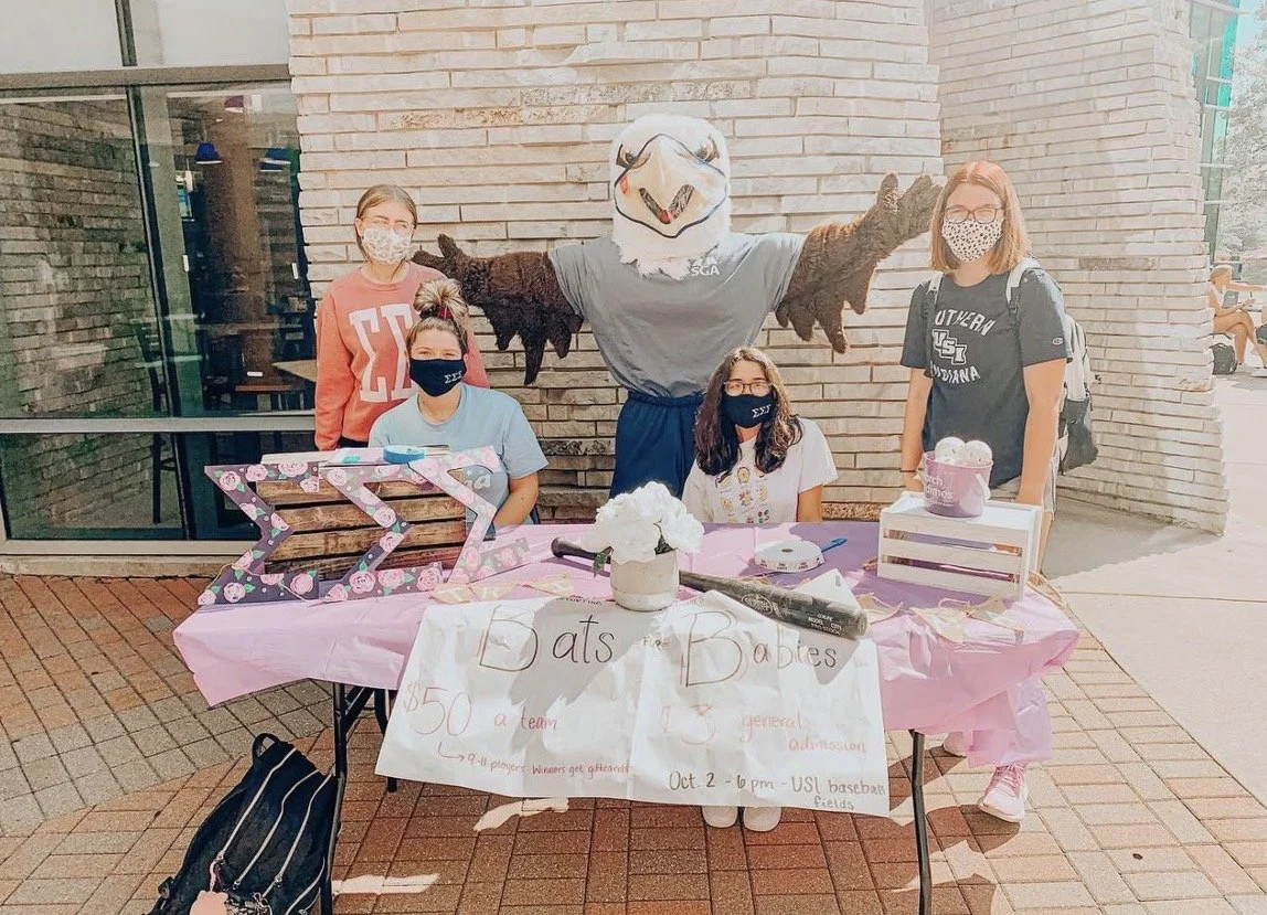 A group of five women, all wearing masks, standing behind a table with pink decorations and signs for a Bake and Babies event at a college campus. One woman is dressed in a bird mascot costume with outstretched arms. The table has baked goods, a bask
