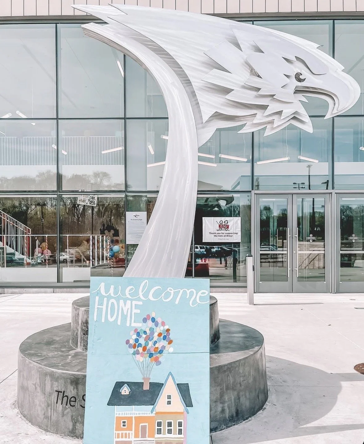 A sculpture of a bird's head with a large curved beak outside a modern glass building. In front of the sculpture, there is a light blue sign with the message 'welcome HOME' and an illustration of a house with balloons above it.