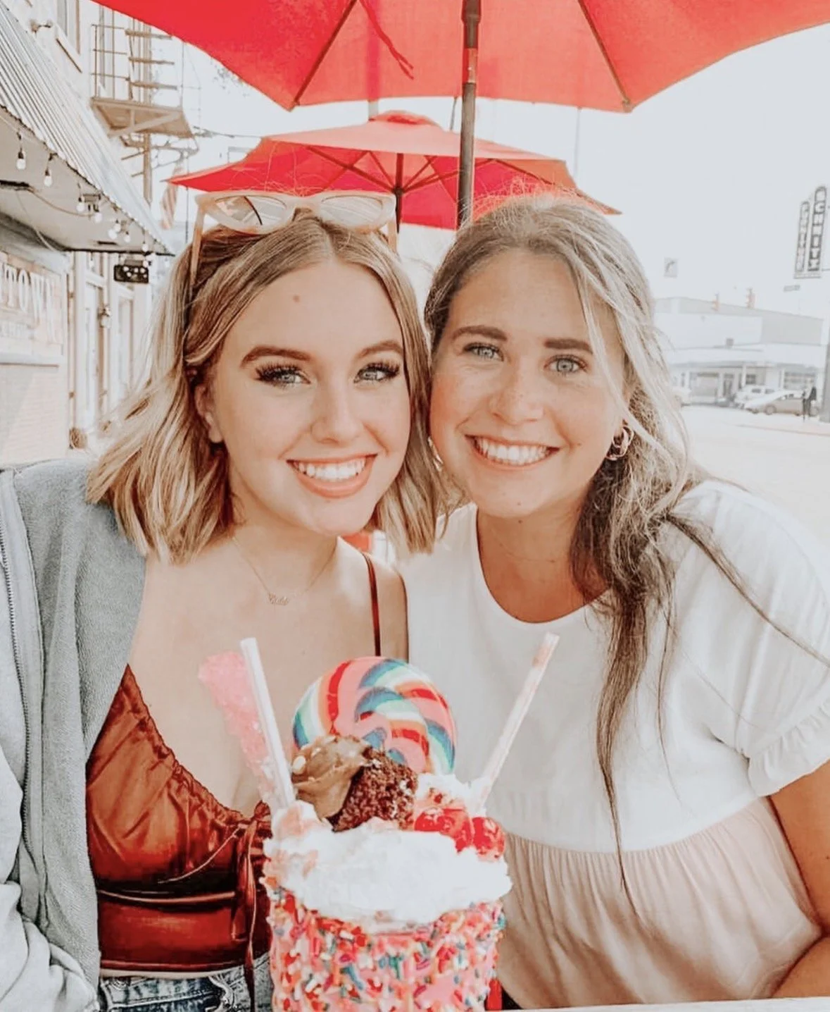 Two smiling women sit outdoors at a cafe table with a large colorful ice cream sundae, garnished with a rainbow lollipop, sprinkles, candy, whipped cream, and two straws. Red umbrellas are visible in the background.