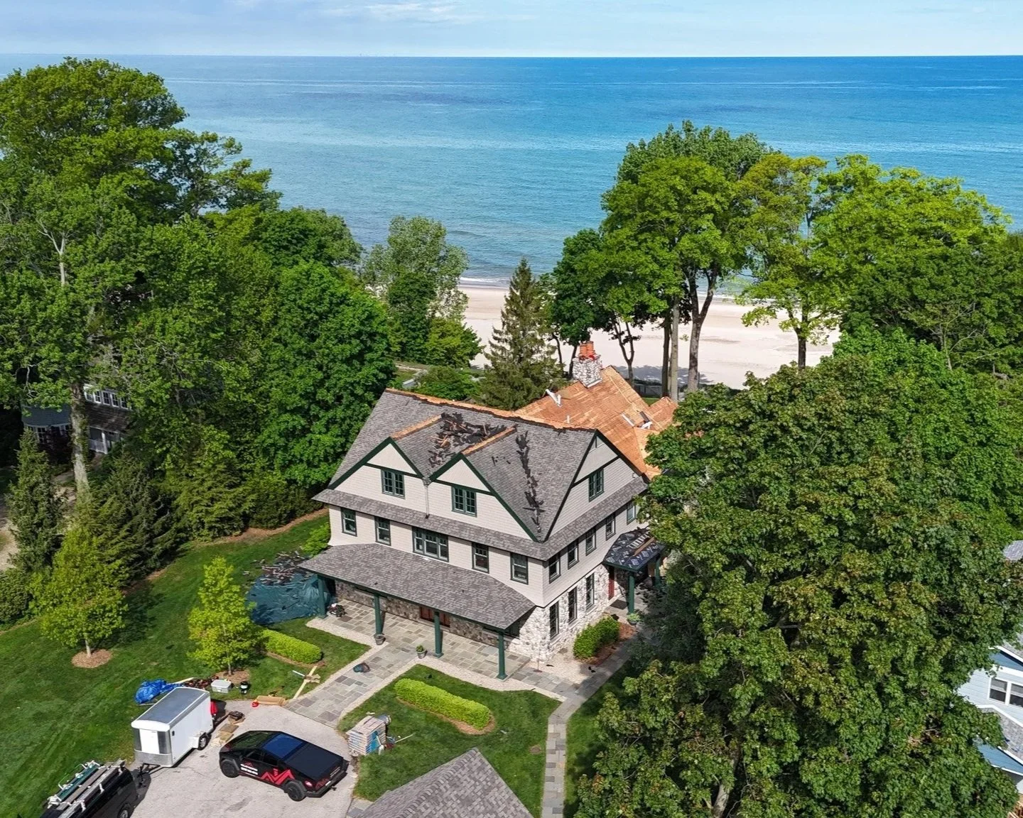 Aerial view of a large house near the beach, surrounded by trees, with parked cars and equipment in the driveway.
