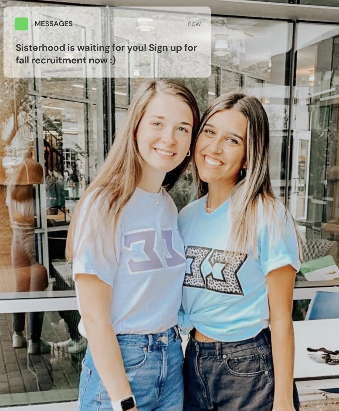 Two women standing inside a building with large glass windows, smiling at the camera. One woman has long light brown hair and is wearing a light blue shirt and jeans, while the other has long dark blonde hair and is wearing a tie-dye shirt with Greek