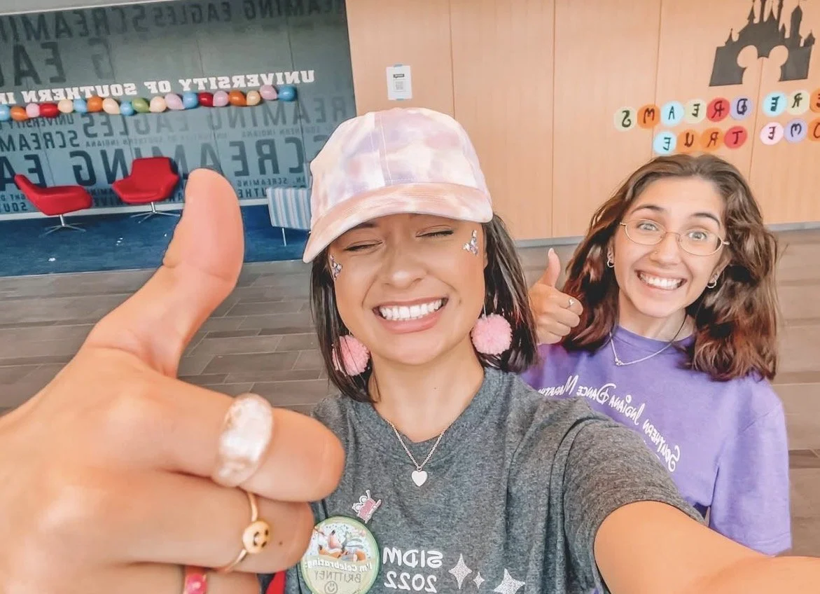 Two young women smiling for a selfie in an indoor setting, one giving a thumbs up. Background features colorful balloons or decorations and words on the wall.