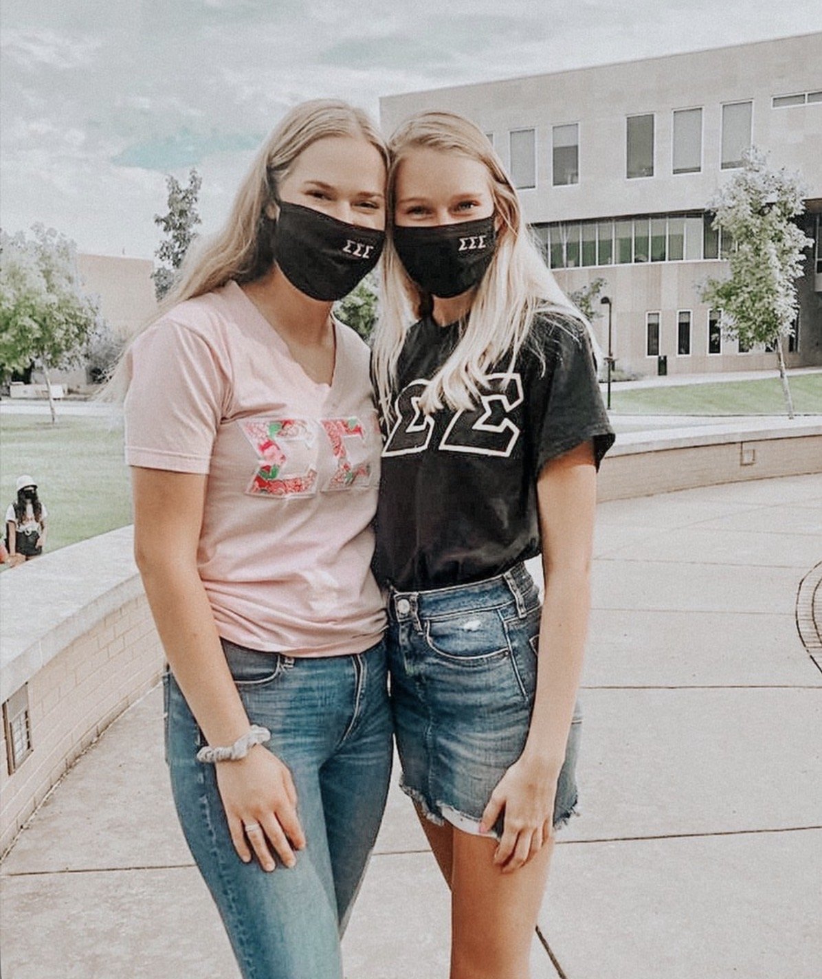 Two young women with blonde hair wearing black face masks, standing outdoors on a college campus. One is wearing a pink shirt with Greek letters, and the other a black shirt with Greek letters, both in casual jeans.