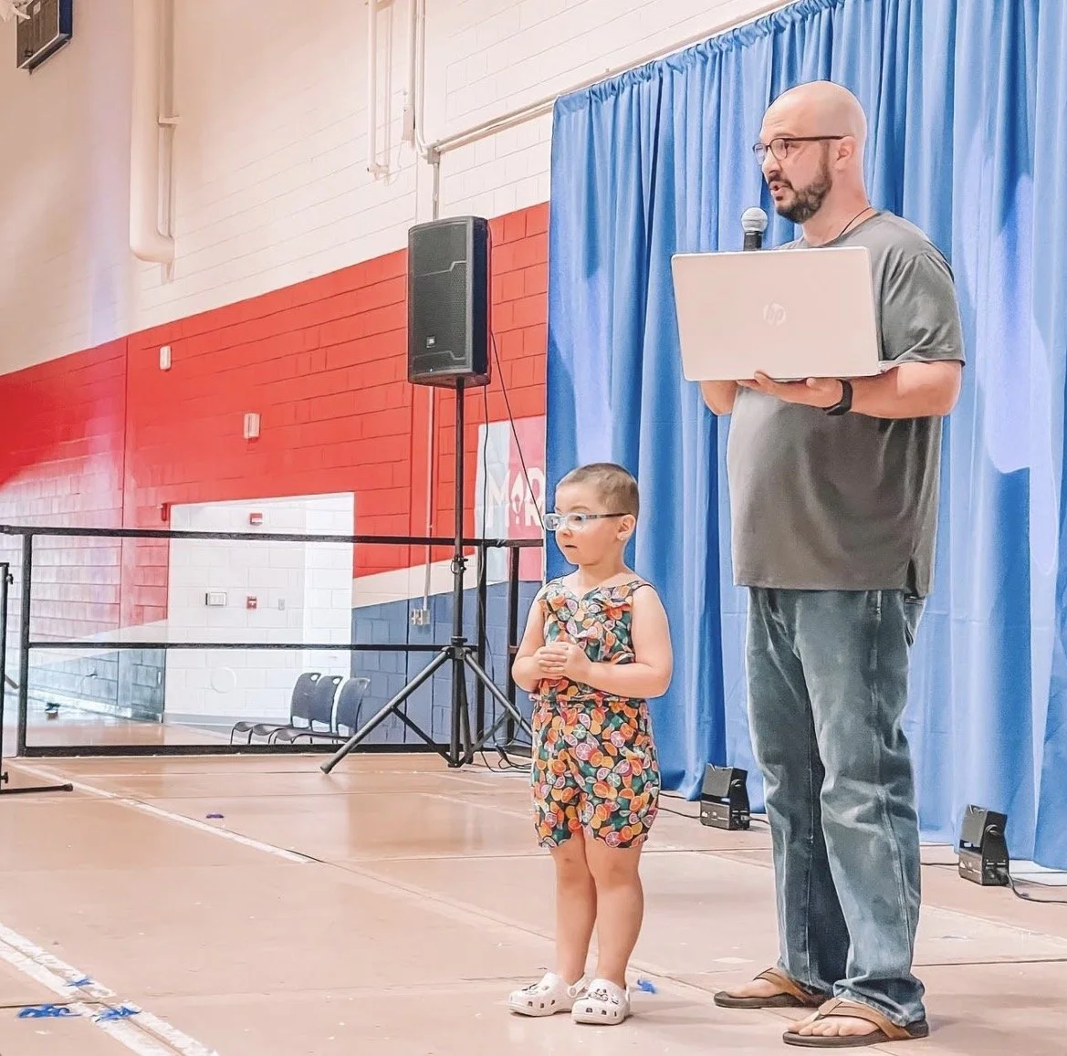 A man and young girl standing on a stage with a blue curtain and speaker behind them, man holding a microphone and a laptop, girl wearing glasses and colorful dress, in a gymnasium with red and white walls.