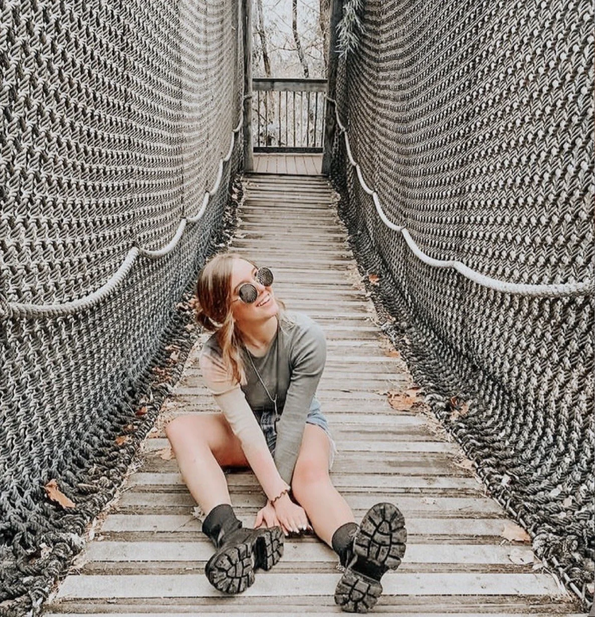 A woman sitting on a wooden bridge, smiling, wearing sunglasses, a long-sleeve shirt, shorts, and hiking boots, surrounded by woven rope sides.