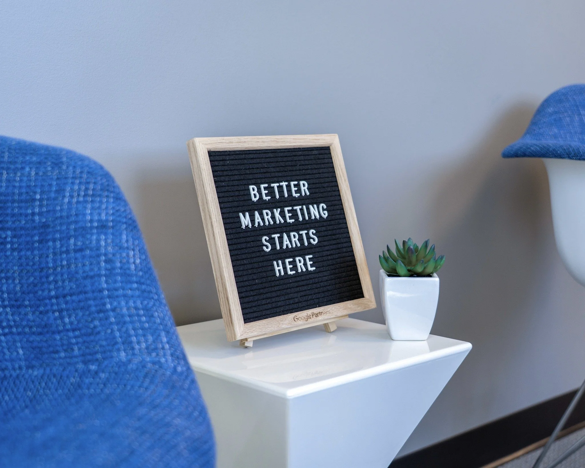 Small black letter board on a white table with the message 'Better Marketing Starts Here', next to a small potted succulent plant.