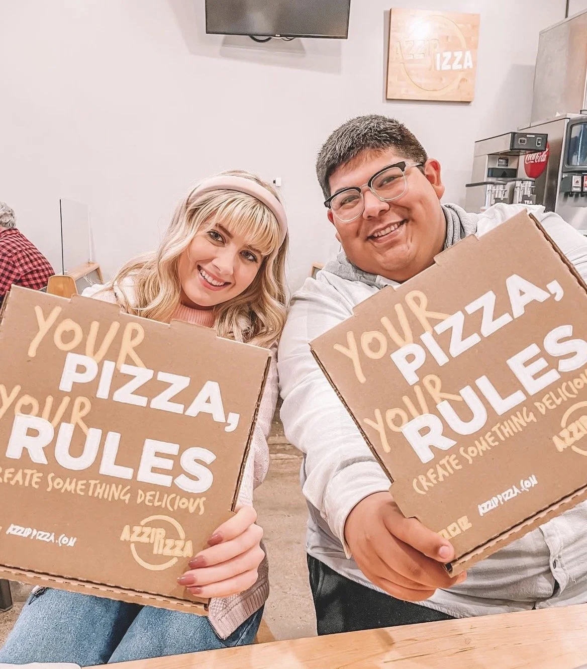 Two people sitting at a table holding pizza boxes with the words 'Your Pizza, Rules' printed on them. The woman has blonde hair and is wearing a pink headband, and the man has short brown hair and glasses. They are smiling inside a pizzeria or casual