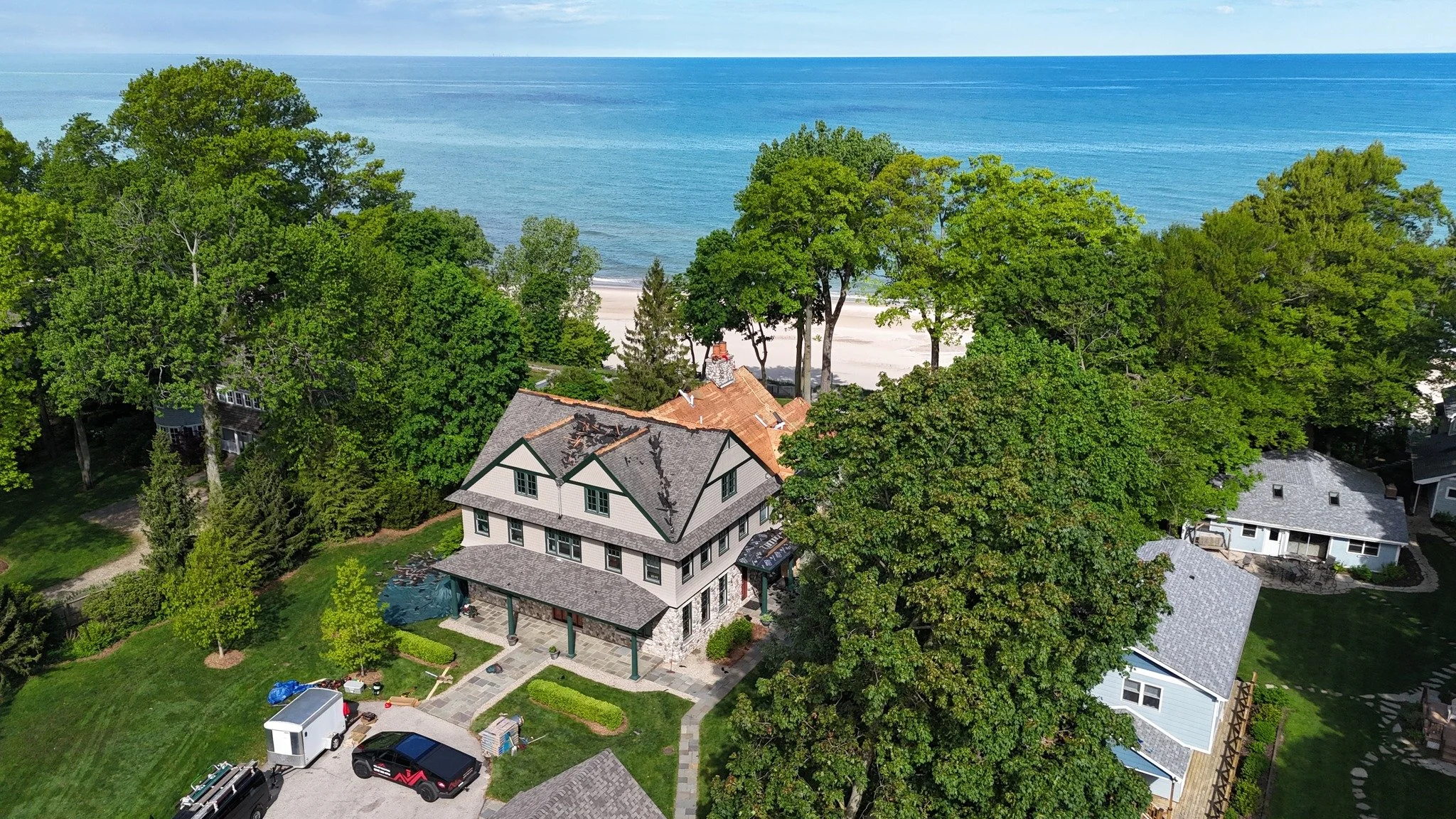 Aerial view of a coastal neighborhood with houses surrounded by trees and a beach with sand and water in the background.