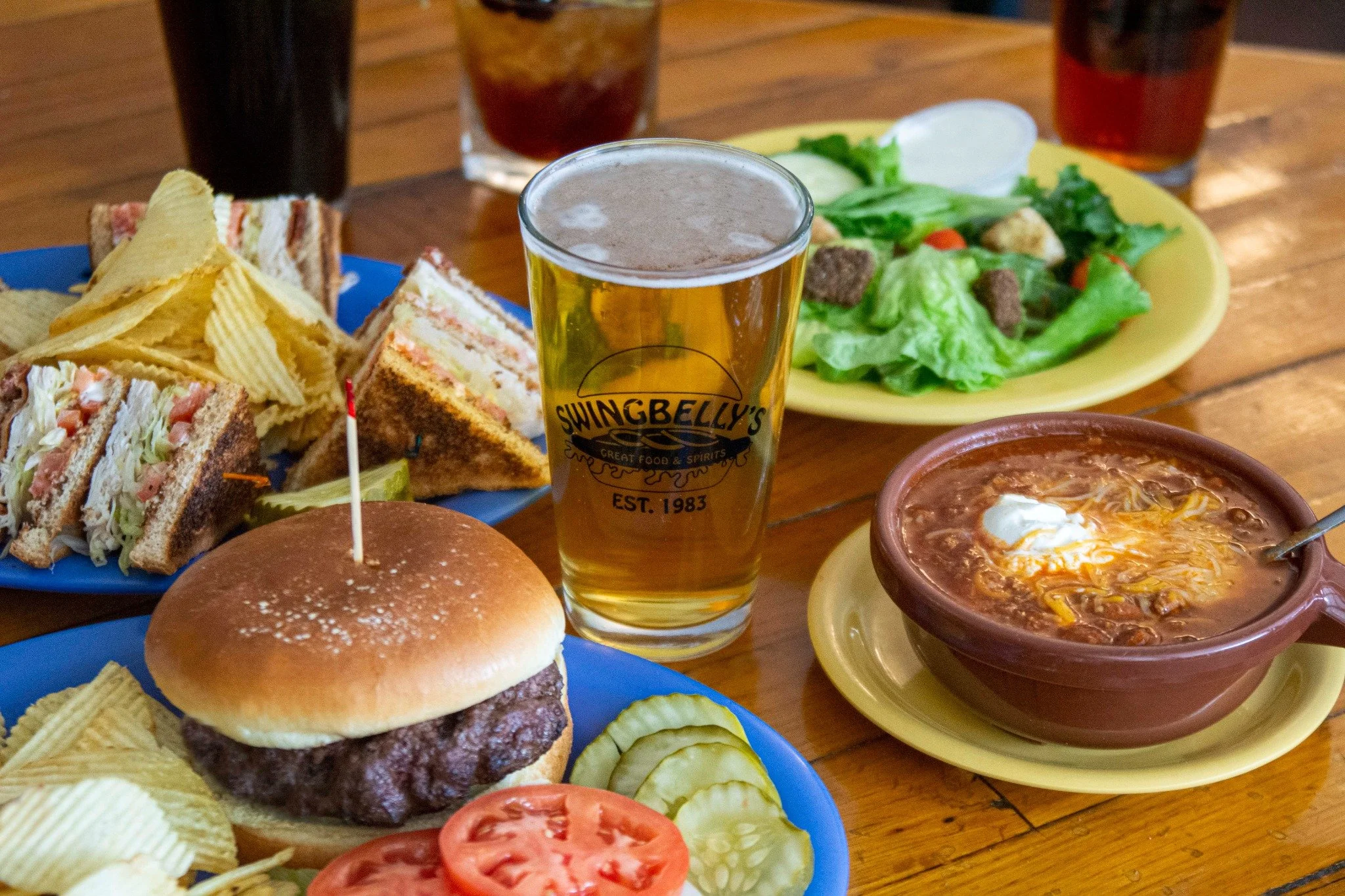A table with a burger, potato chips, deli sandwiches, a salad, a bowl of chili with cheese and sour cream, and a glass of beer at a restaurant.