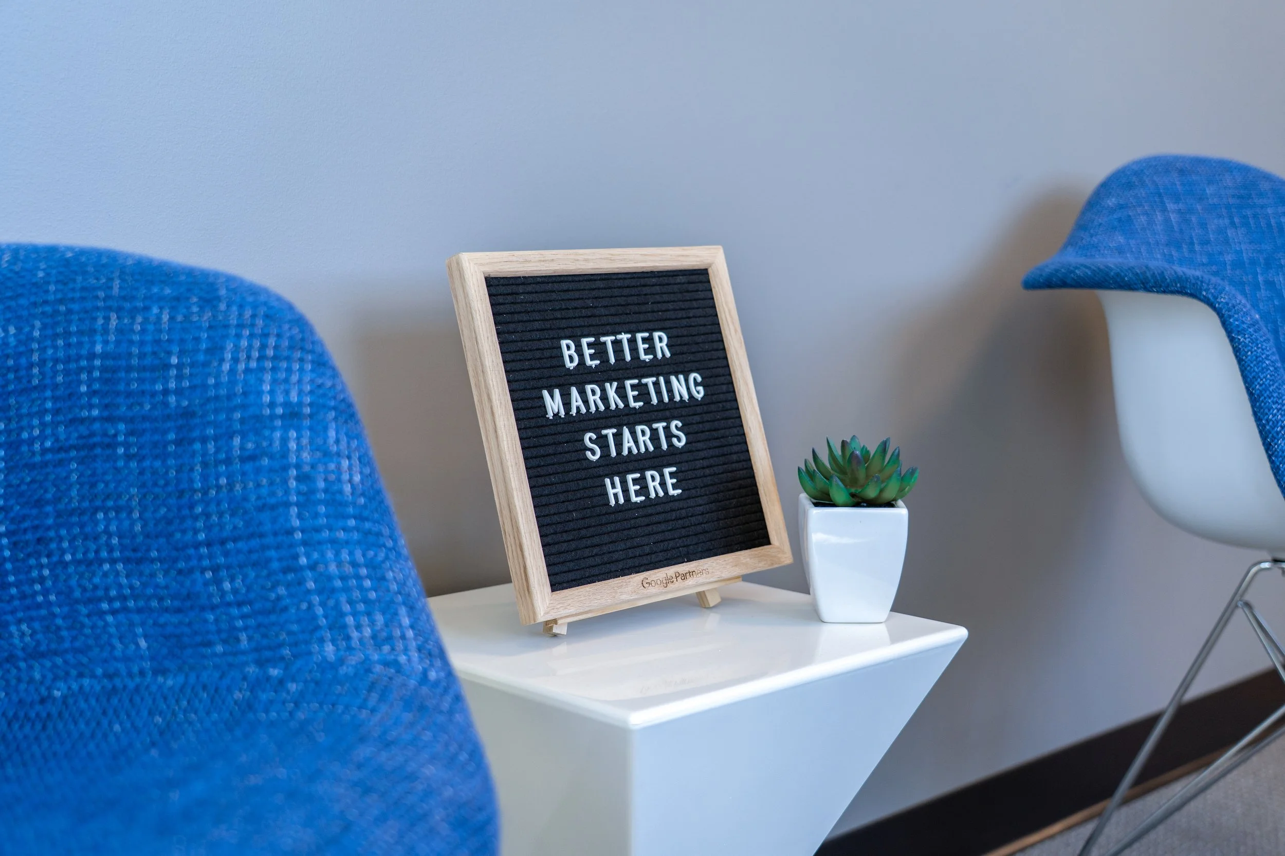 A small black letter board with a wooden frame on a white table, displaying the message 'Better marketing starts here'. Next to it is a small green succulent plant in a white pot. The background shows part of a blue chair and a white wall.