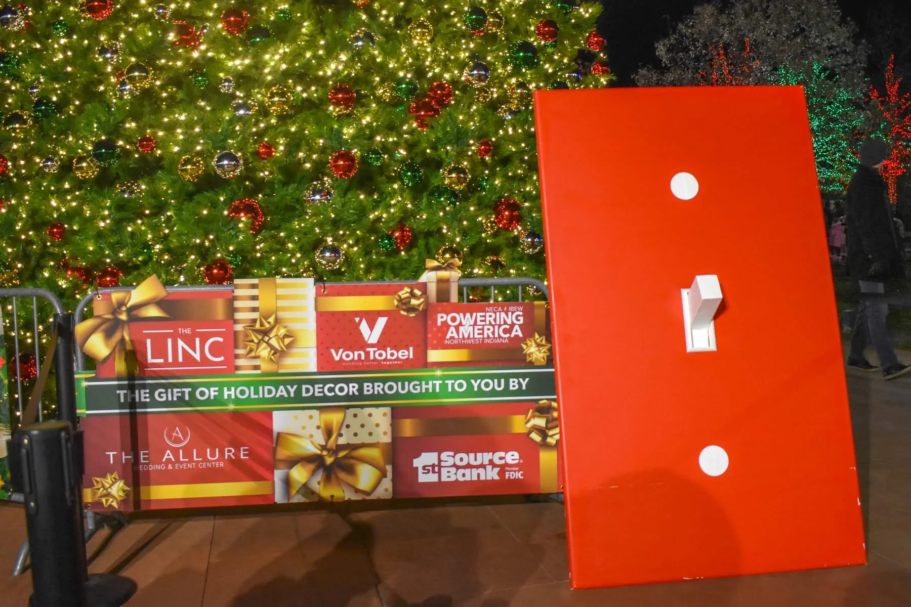 A decorated Christmas tree with lights and ornaments in the background, and a large red donation box with a white slot in the foreground, along with signs and advertisements for local businesses.