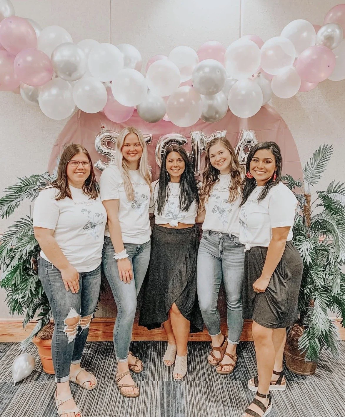 Five women smiling at a celebration, standing in front of a pink backdrop with balloons and silver letters spelling 'SCHOOL'. They are dressed casually, wearing white T-shirts and jeans or skirts, surrounded by potted plants.