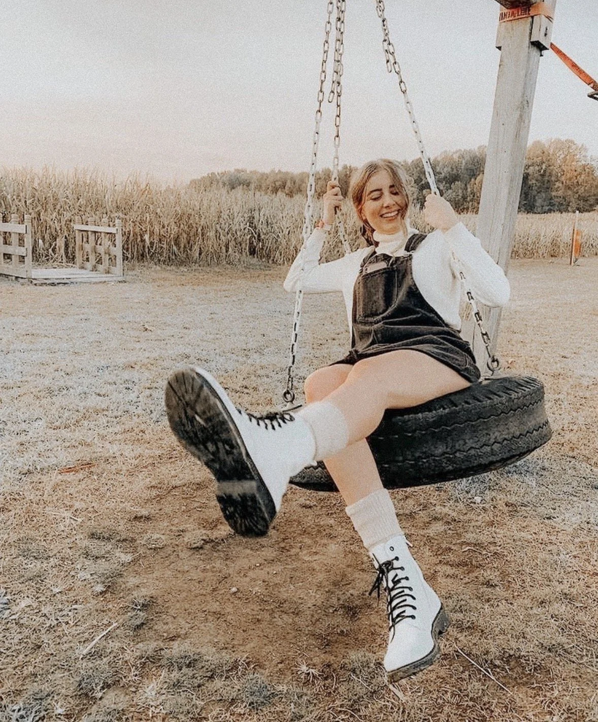 A young woman smiling and sitting on a tire swing in an outdoor field during sunset, wearing white socks, black and white boots, a white long-sleeve shirt, and black overalls.