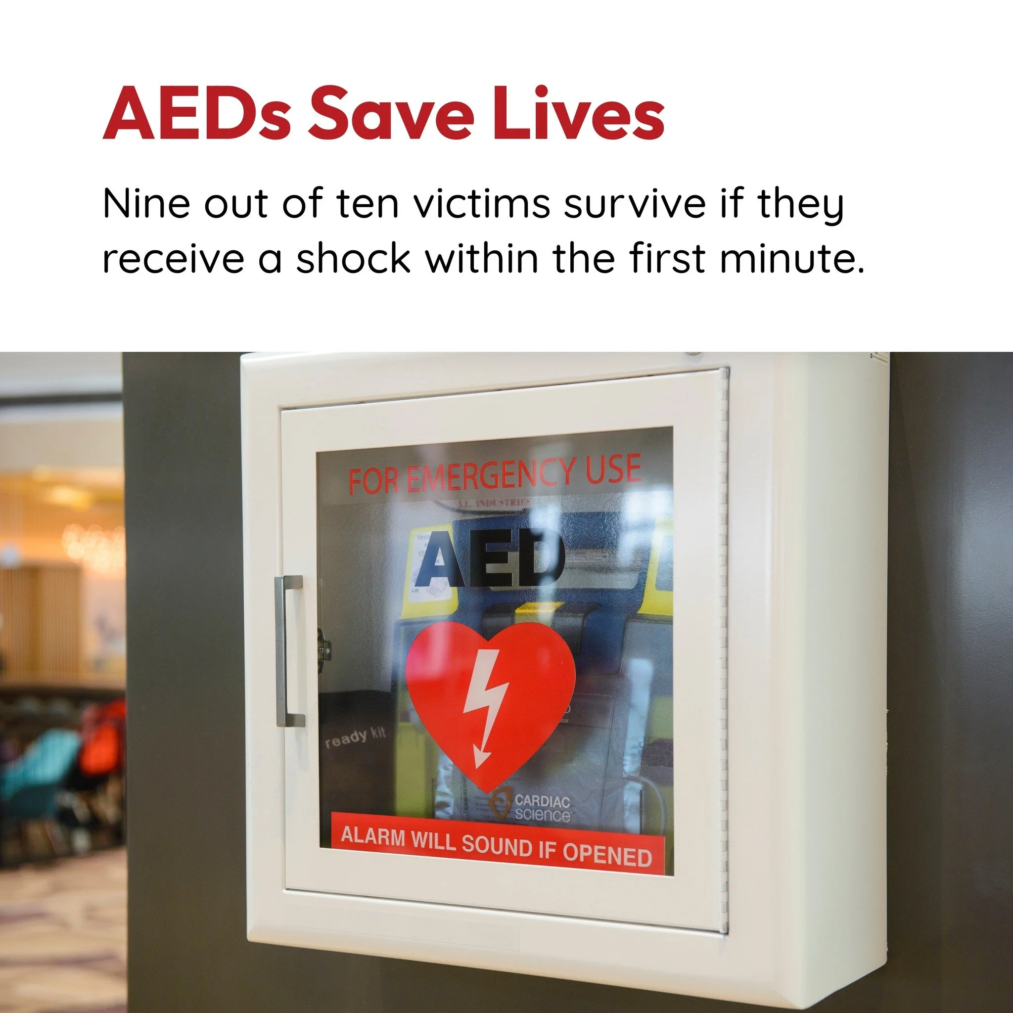 White AED emergency kit cabinet with glass door, red heart symbol with white lightning bolt, and red labels indicating ''FOR EMERGENCY USE'' and ''ALARM WILL SOUND IF OPENED,'' mounted on a wall in a public space.