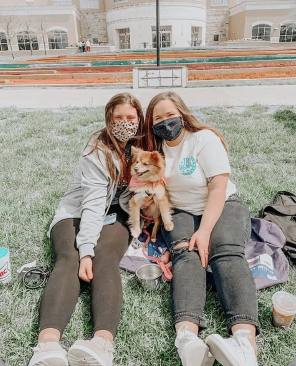 Two young women sitting on grass with a small dog between them, all wearing face masks, in front of a university building. They appear happy, with personal belongings around them.