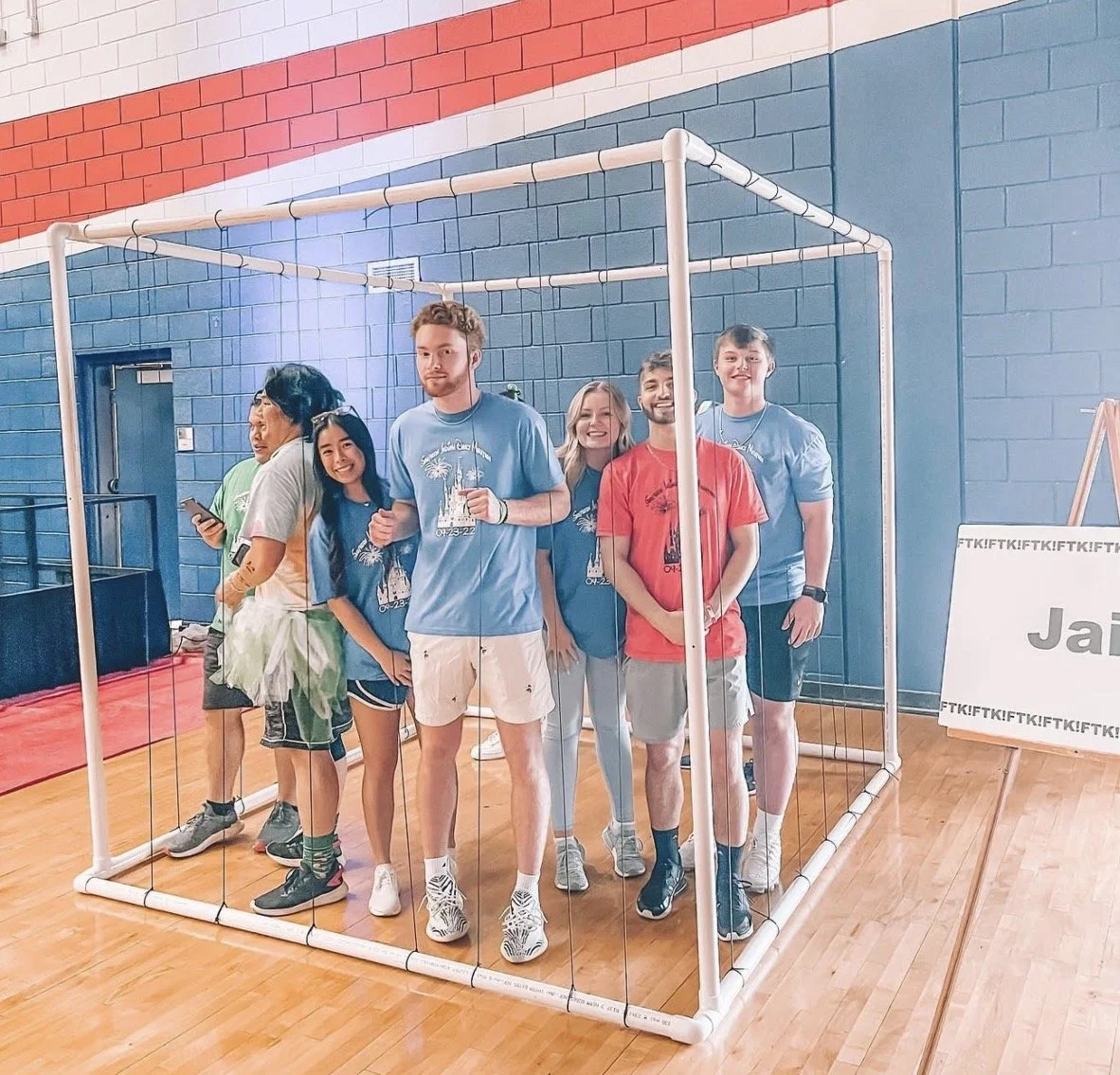 Group of seven young people inside a white wireframe cube, posing for photo at indoor event. Some are smiling, one person is using a phone. Colorful shirts with a design on front, casual athletic shoes, and shorts or casual pants.