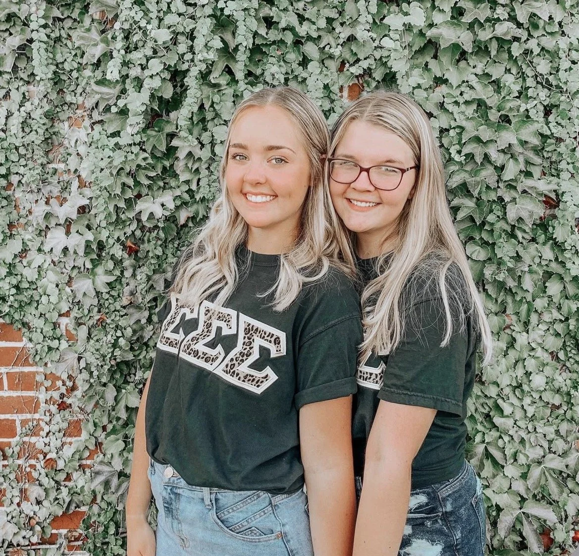 Two young women with long blonde hair smiling and standing close together in front of a leafy green wall. They are wearing black T-shirts.