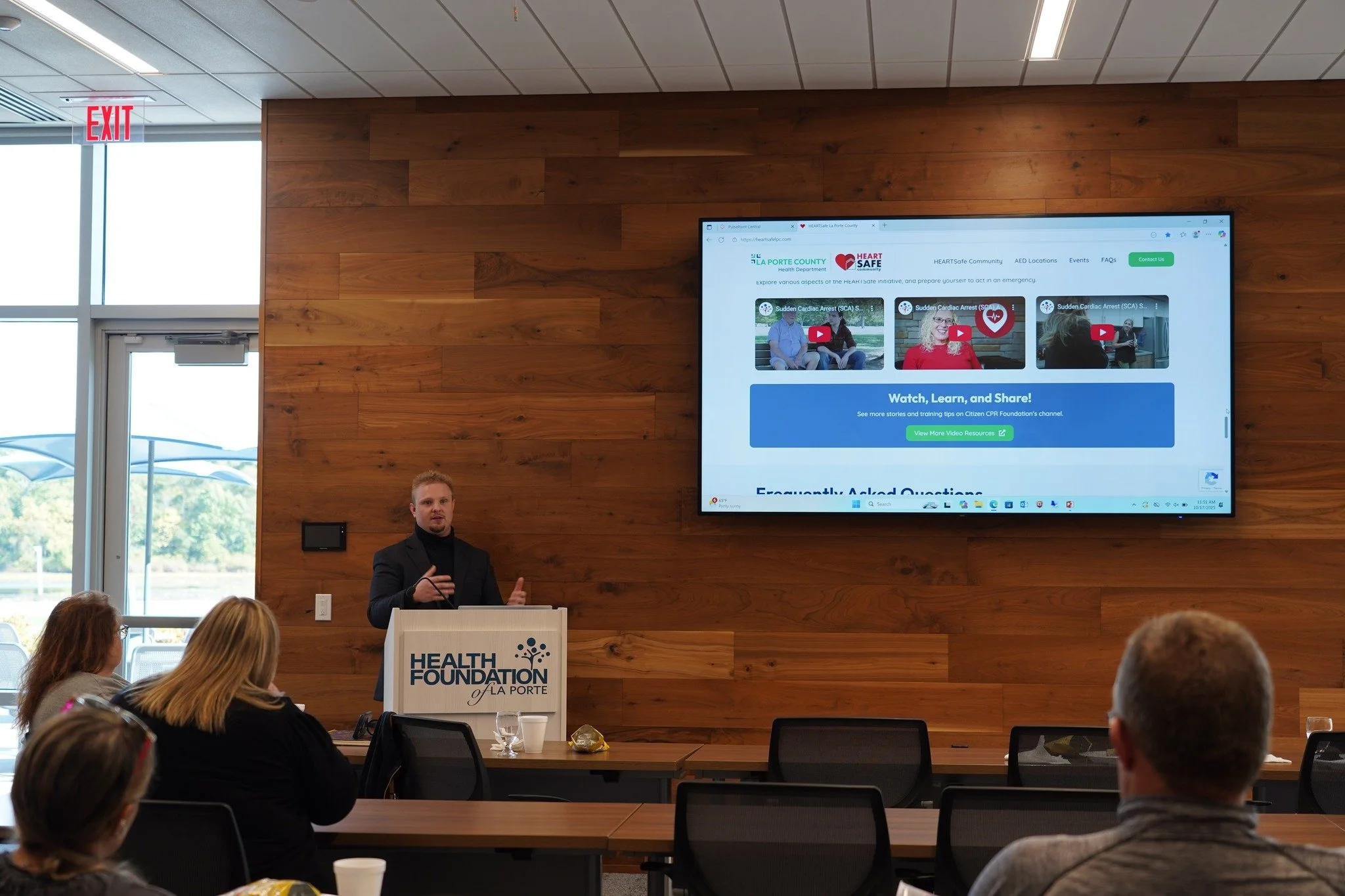 A man giving a presentation at a conference in front of a wooden wall, with a large screen displaying a website about heart safety, while several attendees sit at tables listening.