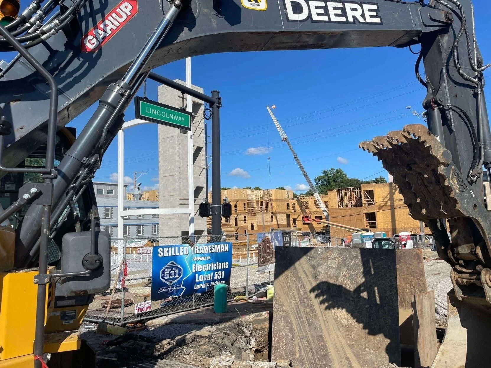 Construction site with a large excavator in the foreground, a crane in the background, and several partially built wooden structures. Street sign reading 'Lincolnway' is visible.