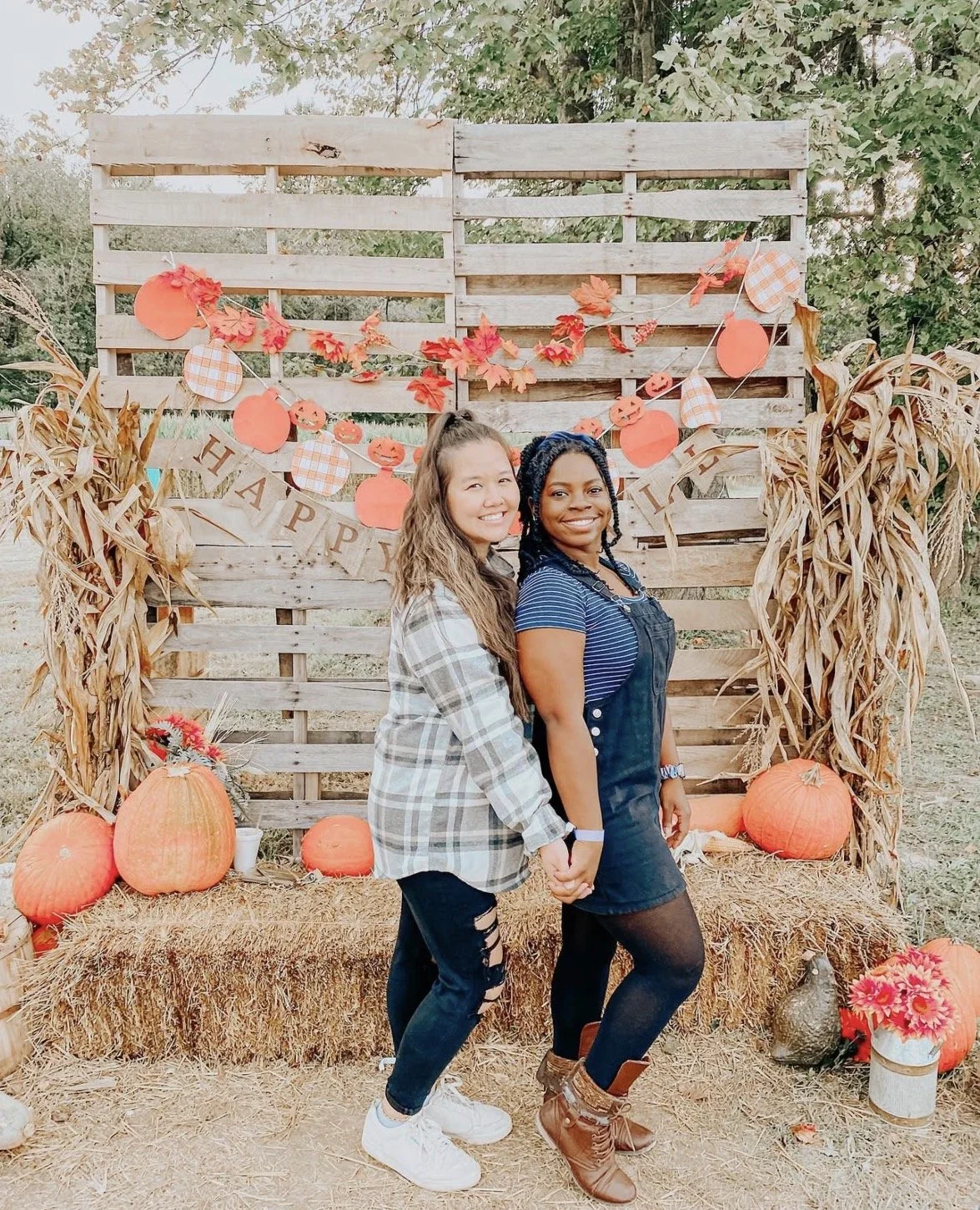 Two women standing outside in front of a wooden fall-themed backdrop decorated with pumpkins, wheat, and paper lanterns, holding hands and smiling.