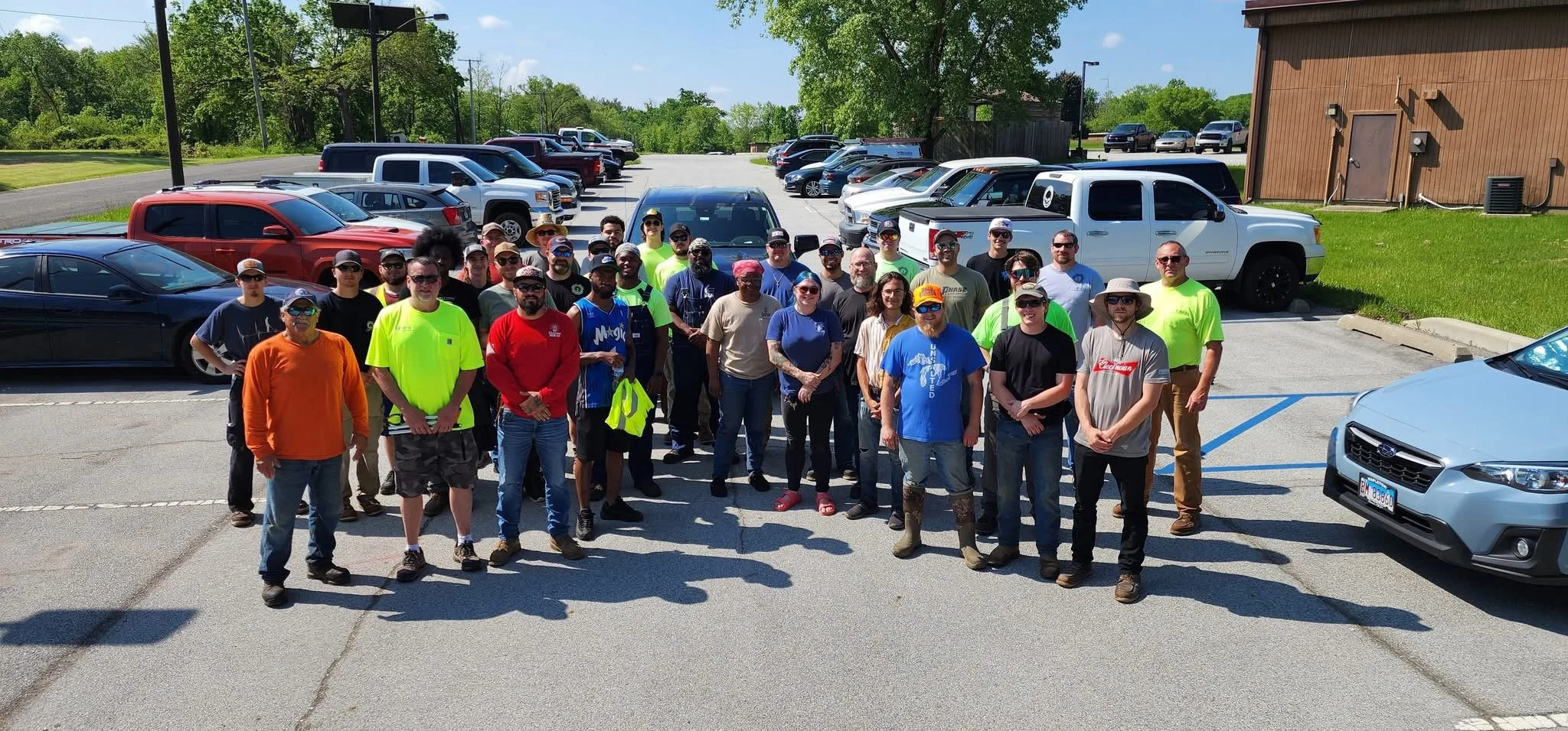 Group of people gathered in a parking lot on a sunny day with a variety of parked cars behind them.