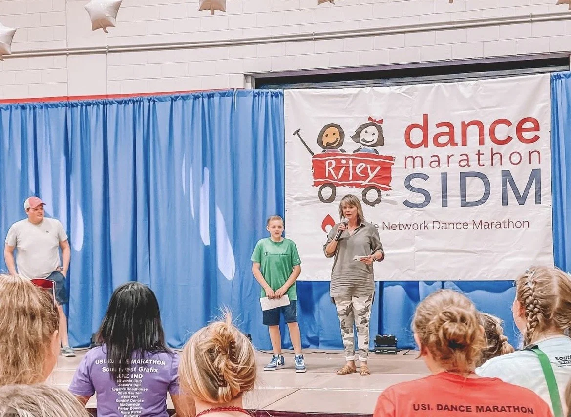 Children and a woman on stage at a dance marathon event, with a large banner behind them that reads 'Riley Dance Marathon' and 'SIDM,' with illustrations of two smiling girls in a red wagon.