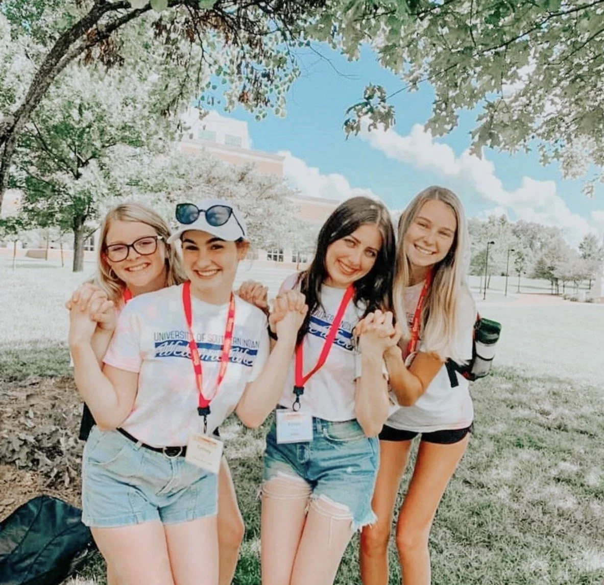 Four young women standing outdoors under trees, smiling, holding hands, wearing casual clothes with university lanyards, on a sunny day with a blue sky and some clouds.