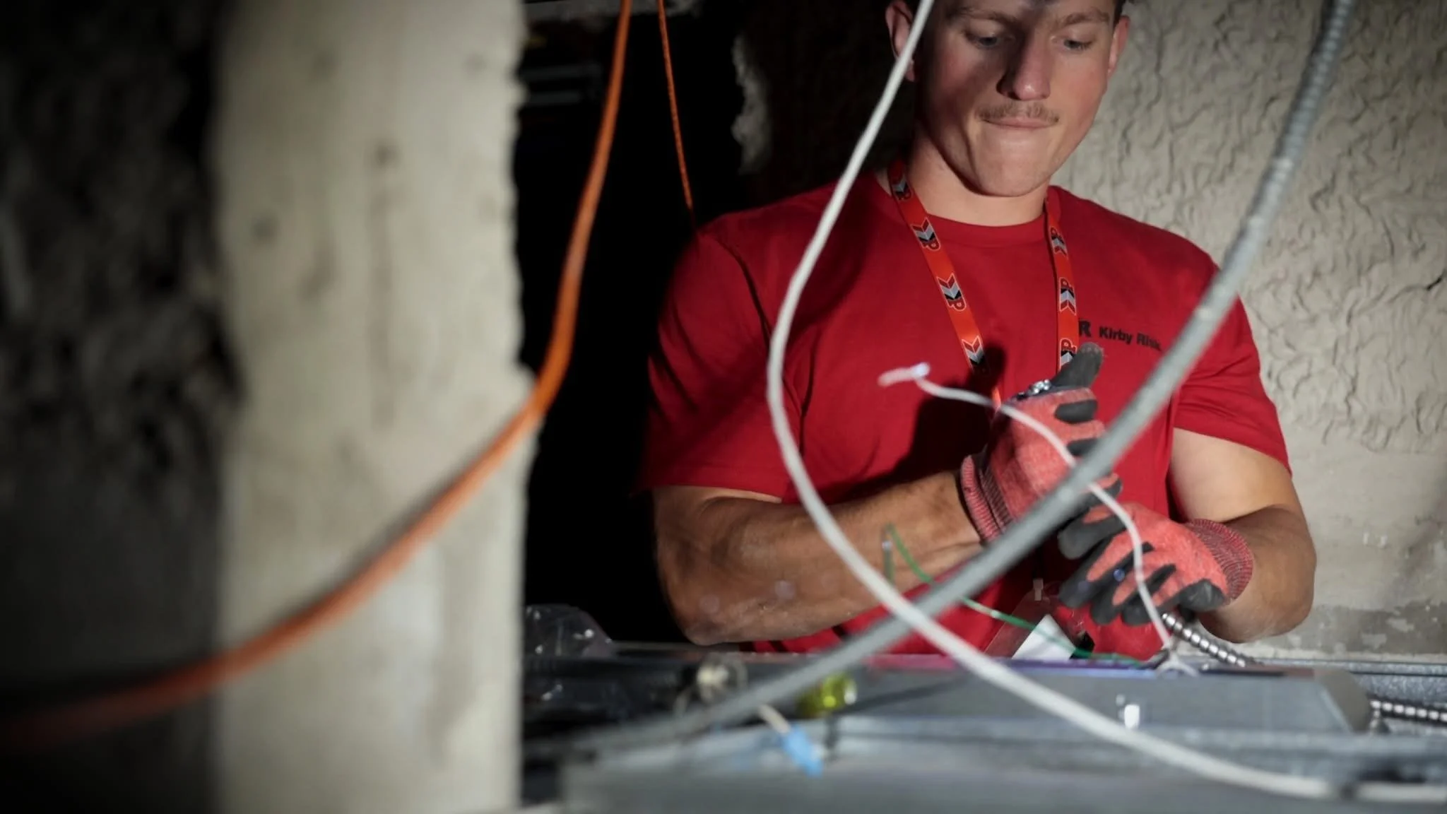A young male technician wearing a red shirt and gloves working with electrical wires inside a wall cavity.