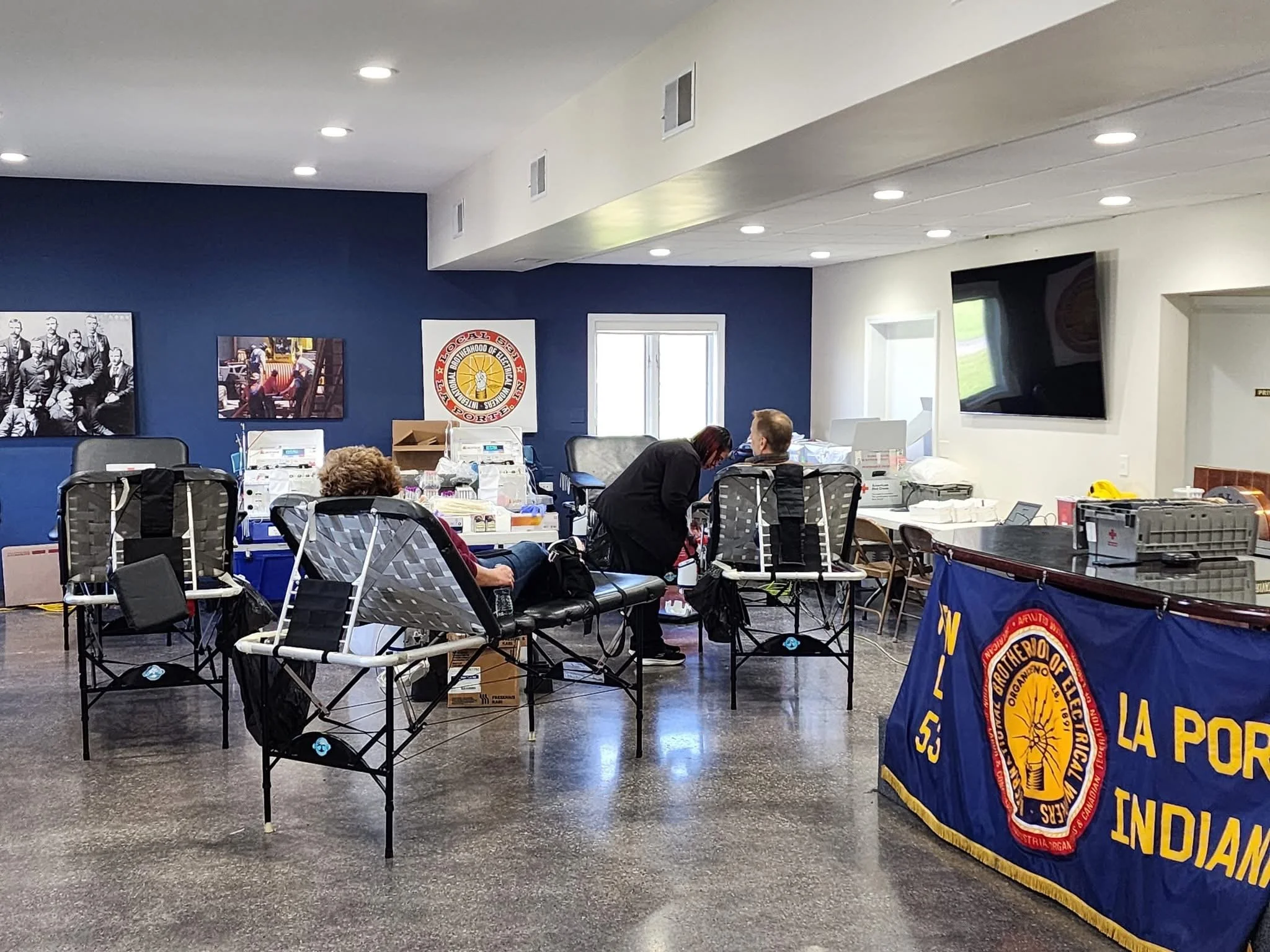 Room with people seated in lawn chairs, receiving blood donations at a community blood drive, with banners and posters on the walls, including a large flag and a logo for the Southern Indiana Fire Department.