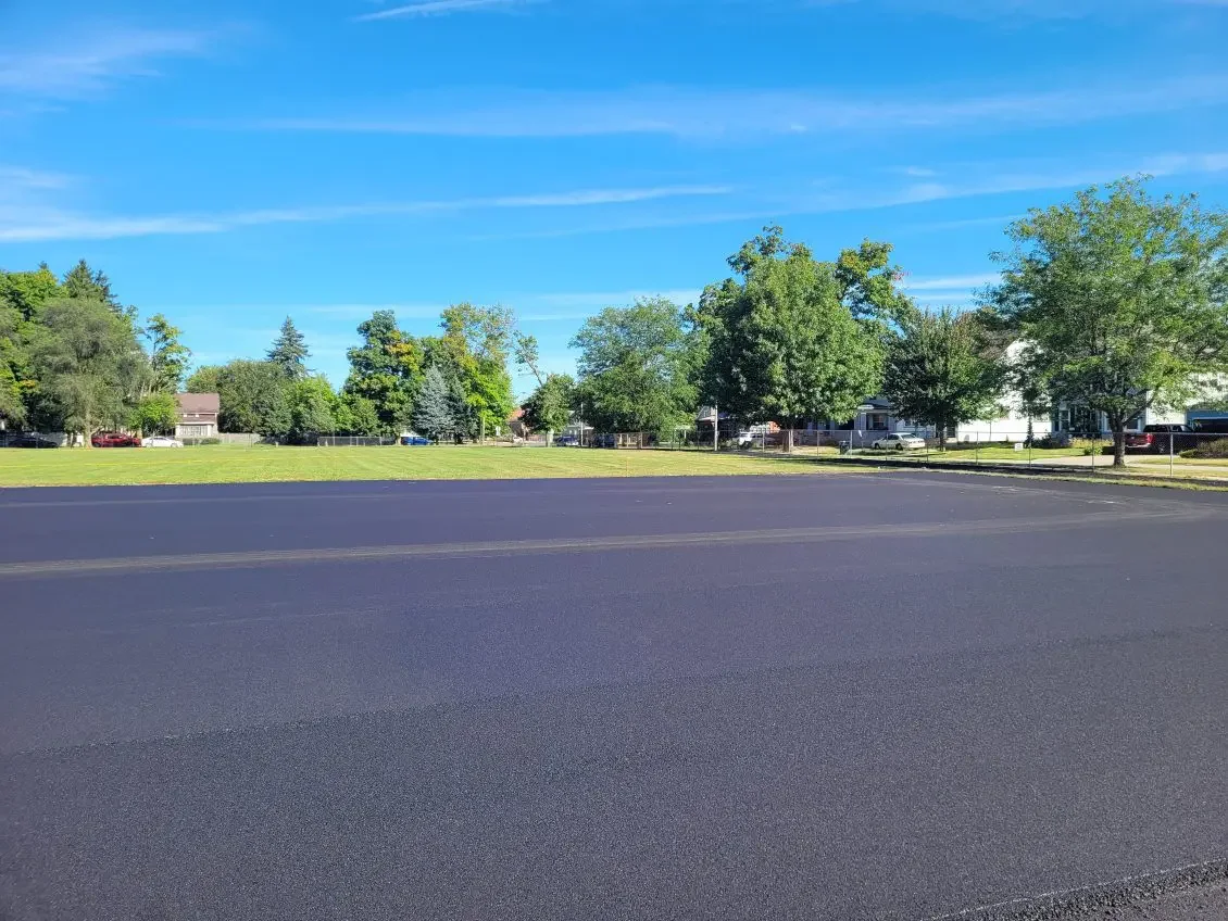 A freshly paved black asphalt road in a suburban neighborhood with green trees, houses, and a clear blue sky.