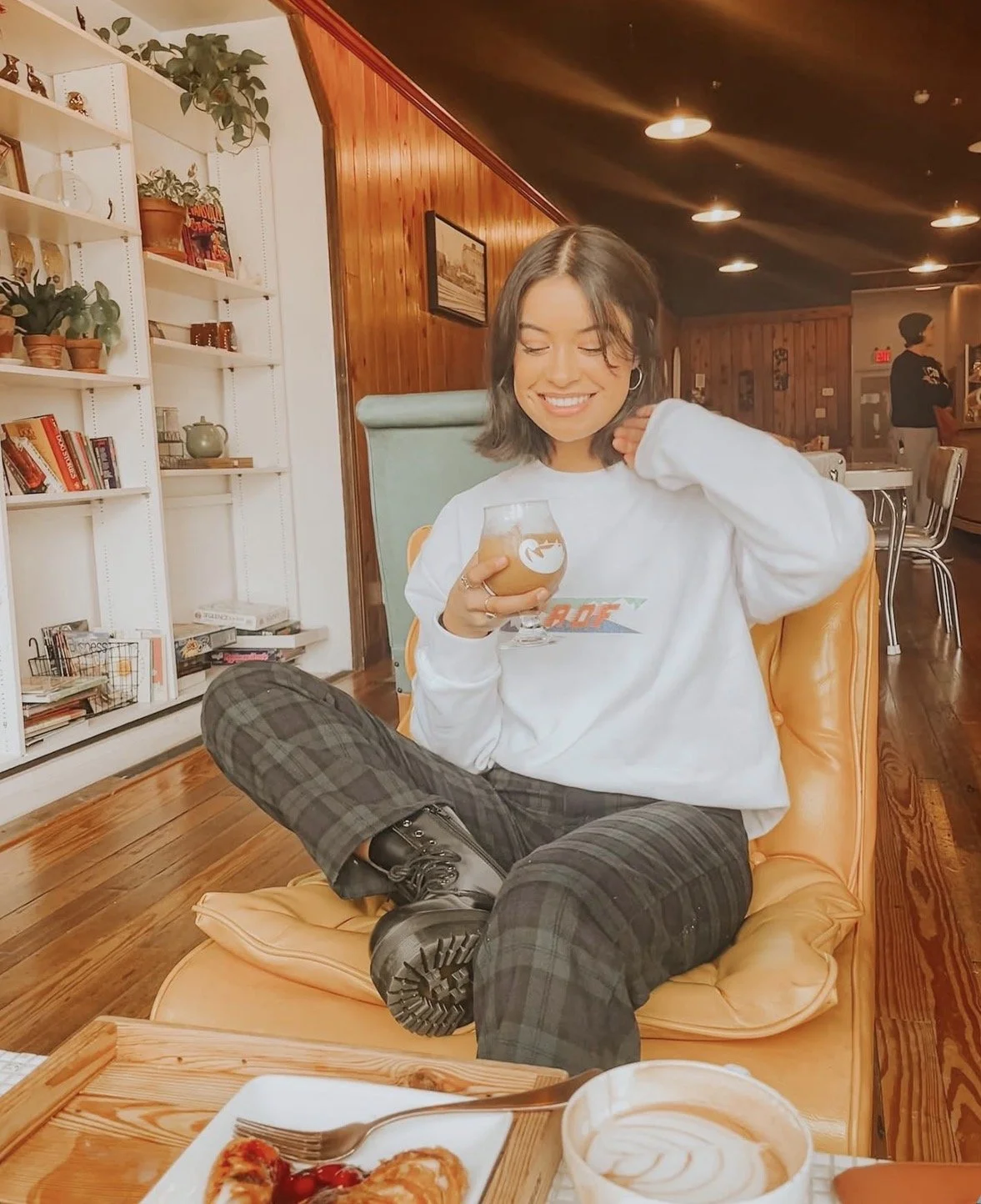 A cheerful young woman with shoulder-length dark hair, wearing a white sweatshirt and plaid pants, sitting cross-legged on a yellow lounge chair, holding a coffee drink, in a cozy cafe with wooden walls and bookshelves.