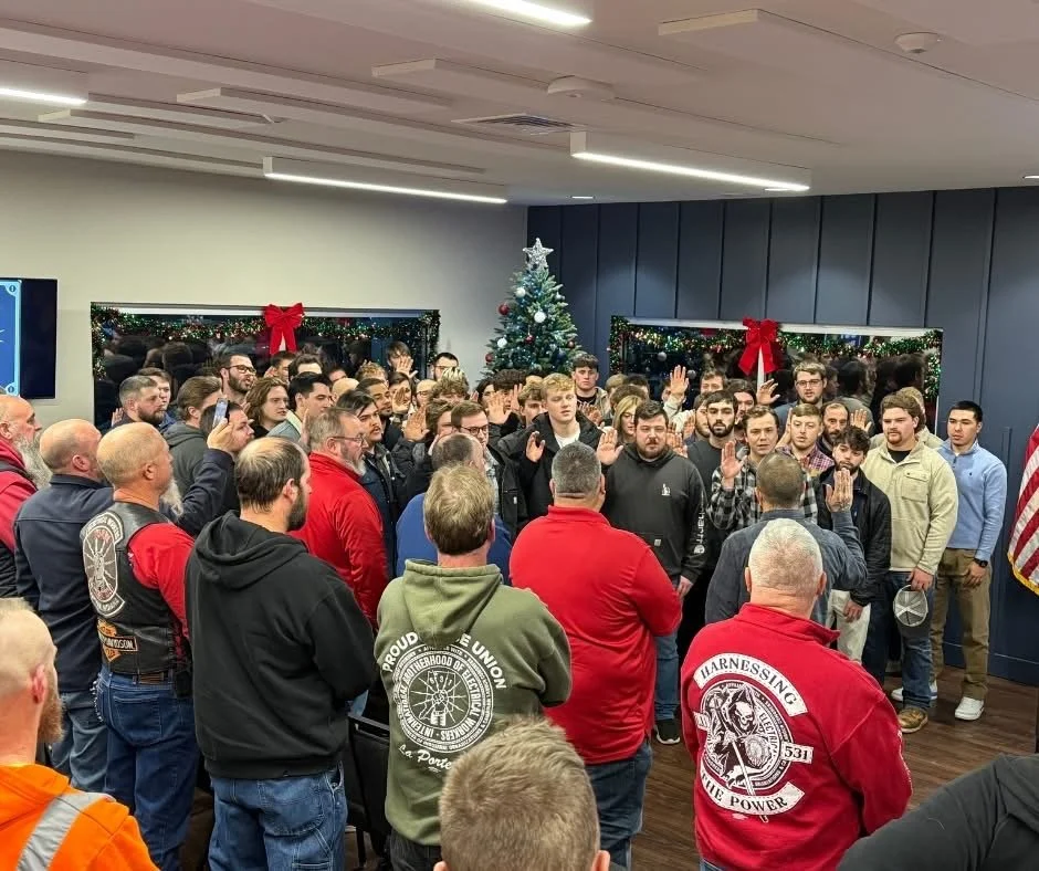 A group of people, mostly young men, gathered in a room decorated for Christmas, with a Christmas tree and holiday decorations, as some raise their right hand during a pledge or oath.
