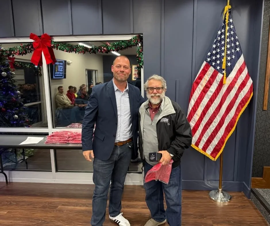 Two men standing indoors near an American flag, decorated for Christmas with a bow and garland, with a Christmas tree and people visible through a window in the background.